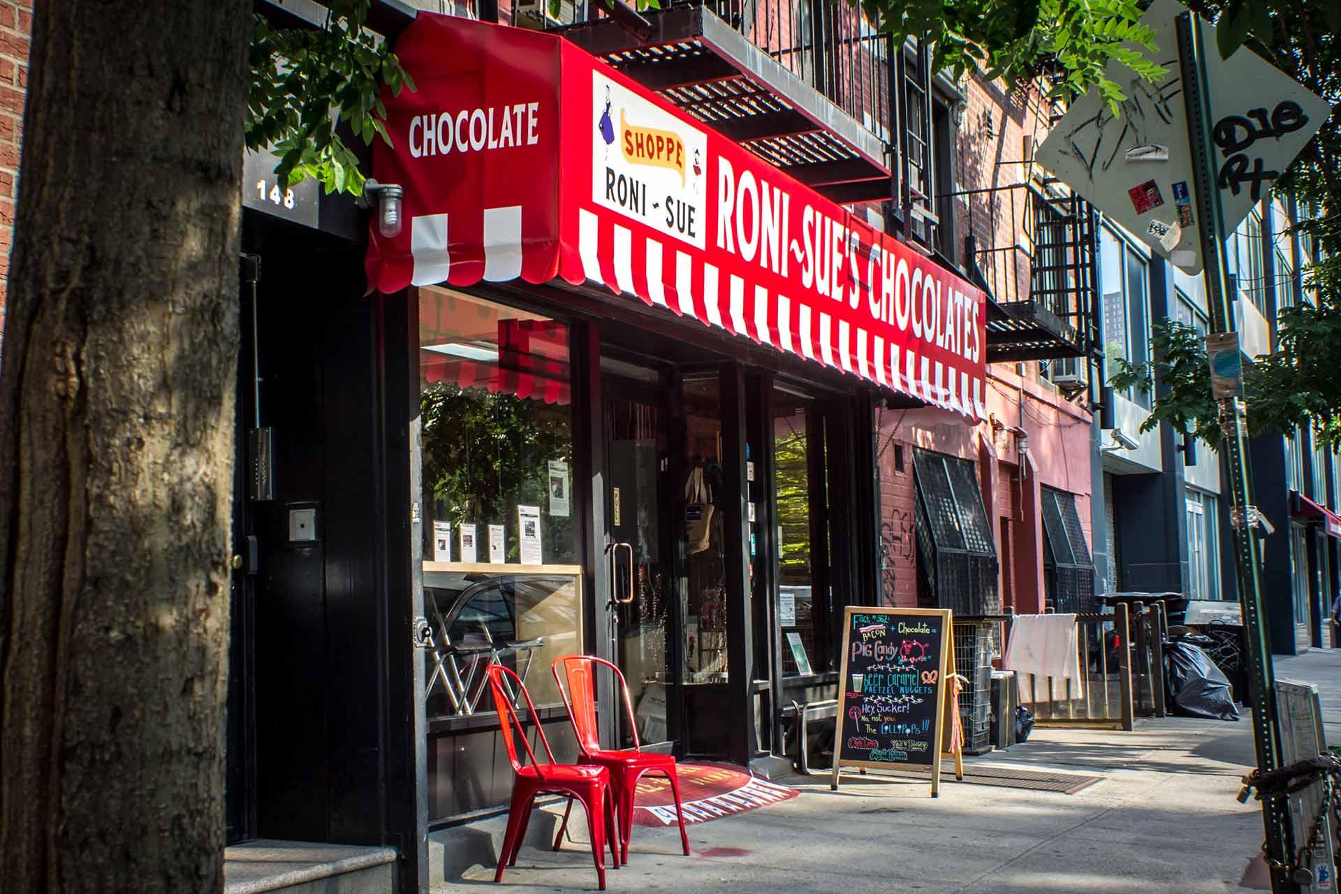 Storefront with a red and white striped awning reading “RONI-SUE'S CHOCOLATE,” two red chairs outside, a chalkboard menu on the sidewalk, and trees and other buildings lining the street.