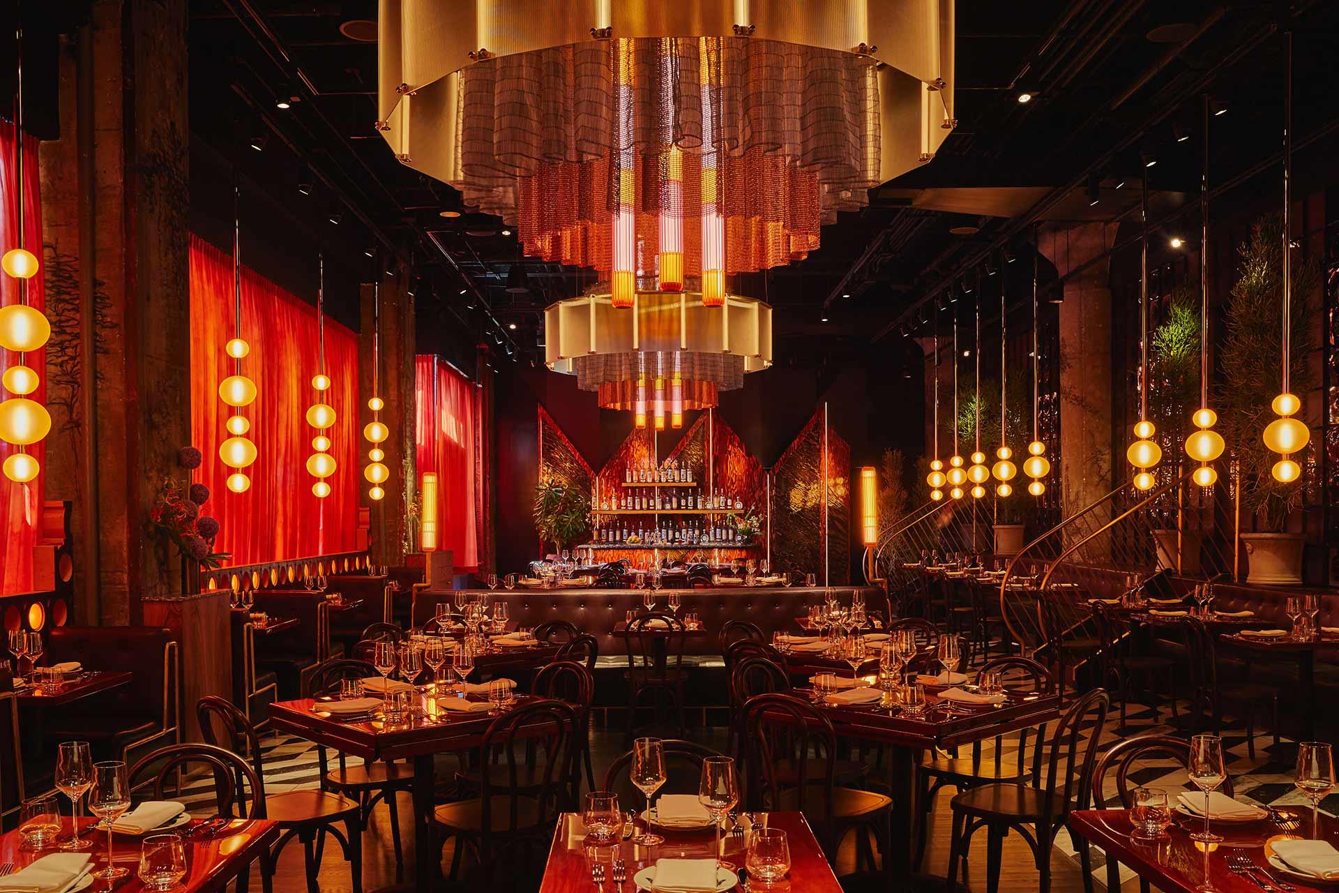A dimly lit, upscale restaurant with elegant chandeliers, red curtains, globe pendant lights, and neatly set wooden tables, creating a warm and inviting ambiance. The bar is illuminated at the far end of the room.