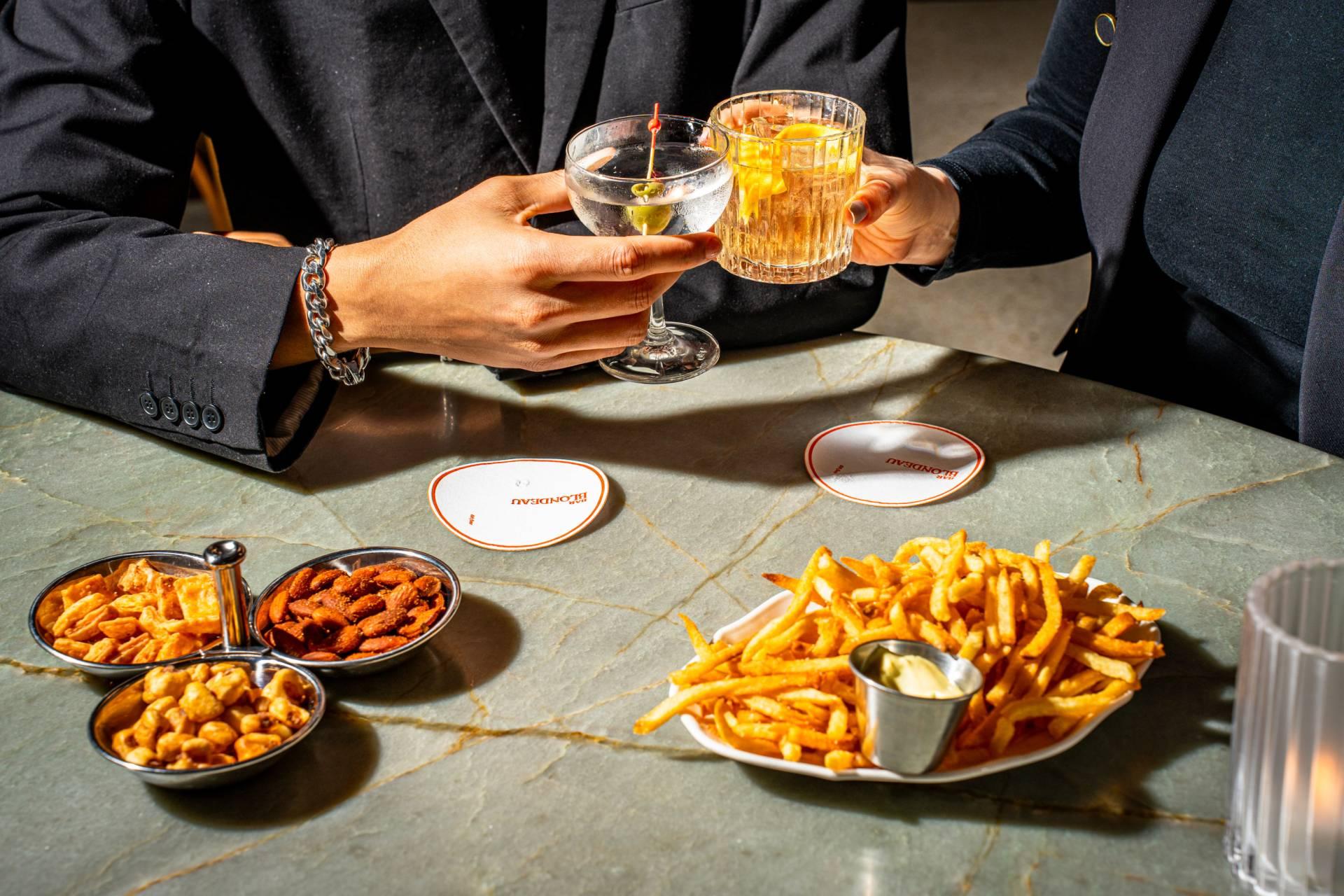 Two people clink glasses over a marble table with French fries, snack mixes, and dips. Both wear dark blazers, and the table is brightly lit with two drink coasters visible.