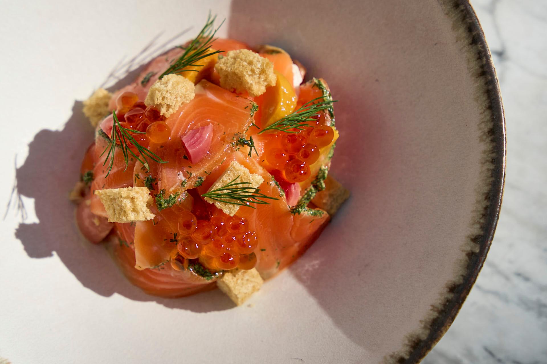 A dish of smoked salmon adorned with red roe, fresh dill sprigs, and small croutons, served in a shallow, white bowl. The background features a marble surface.