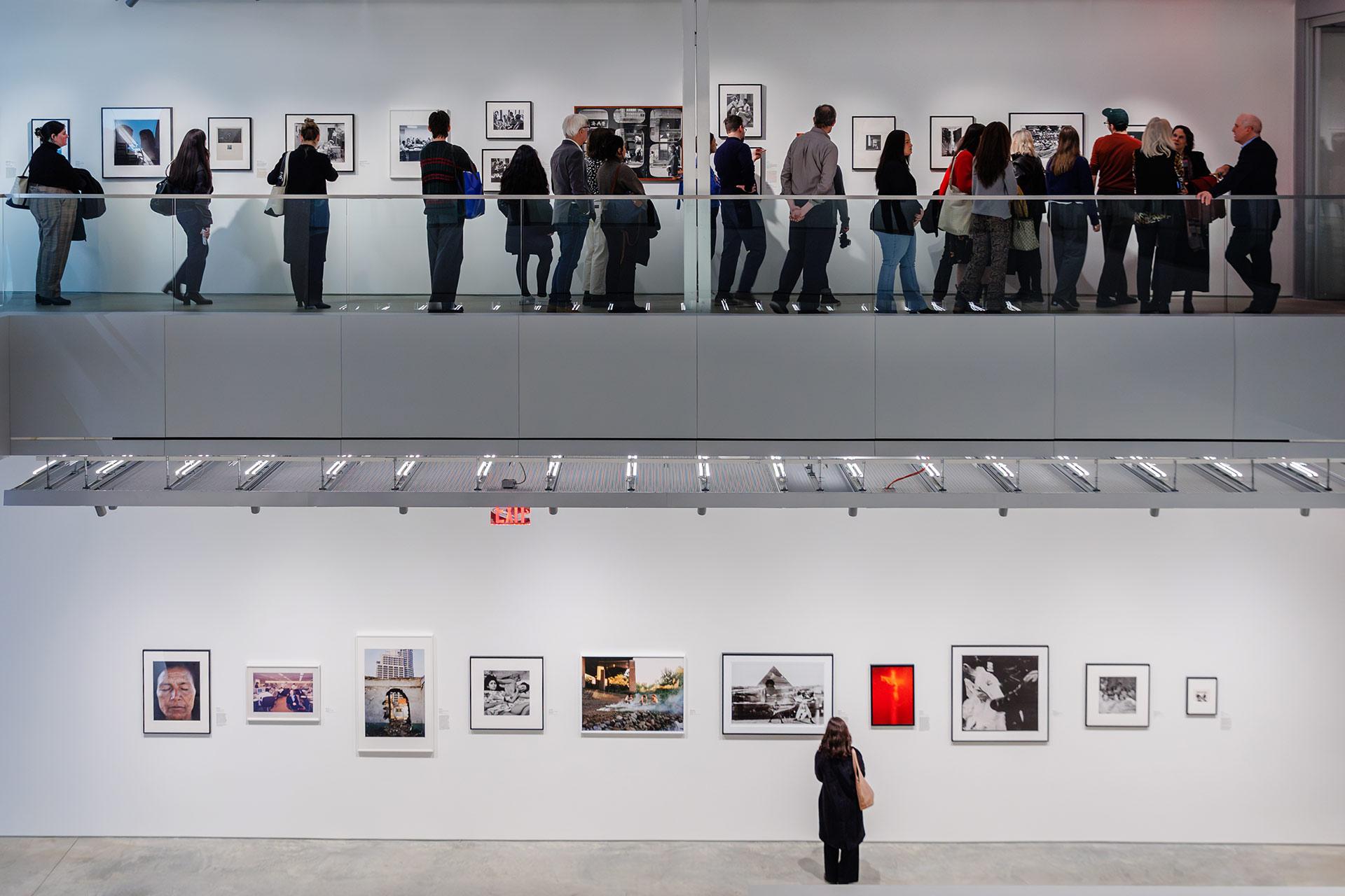 A woman stands alone viewing framed art on a white gallery wall while a group of people looks at artwork on the upper level behind a glass railing. Both walls display various photographs and artworks.