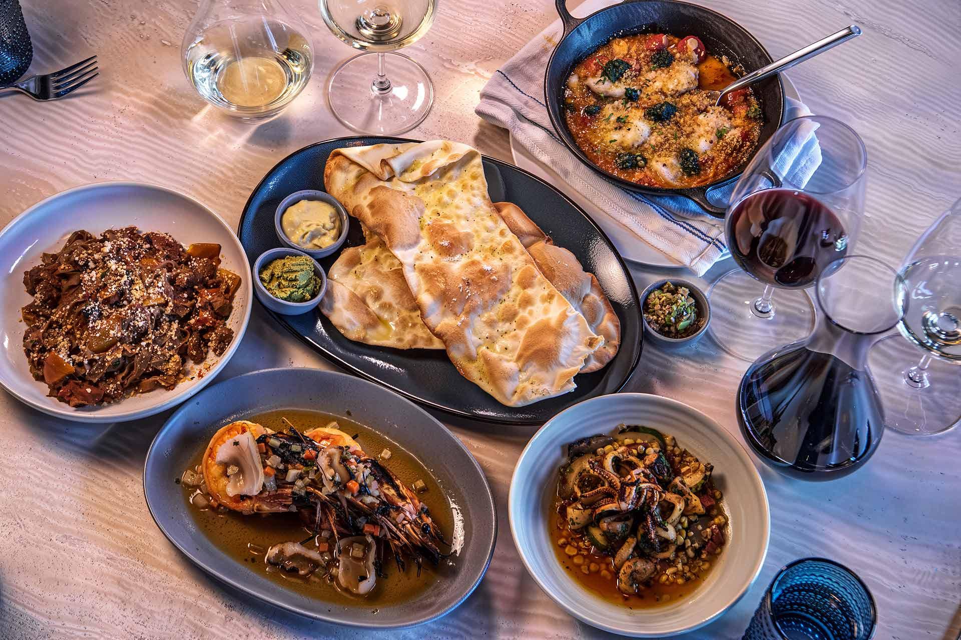 A table set with various Italian dishes, including flatbread, seafood, meat dishes, small bowls of sauces, and glasses of red and white wine on a light wood surface.