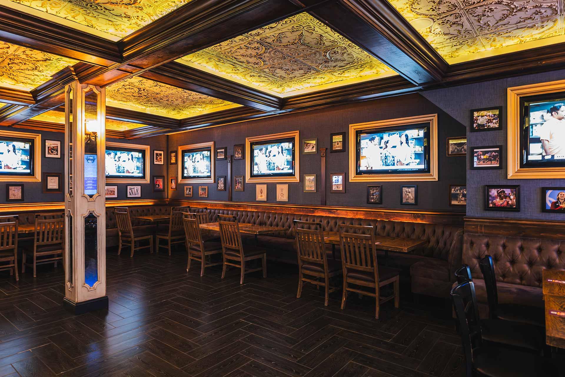 A cozy restaurant interior with tufted booth seating, wooden chairs, framed photos, and multiple TV screens on dark-paneled walls. Warm yellow lighting from decorative ceiling panels creates an inviting atmosphere.
