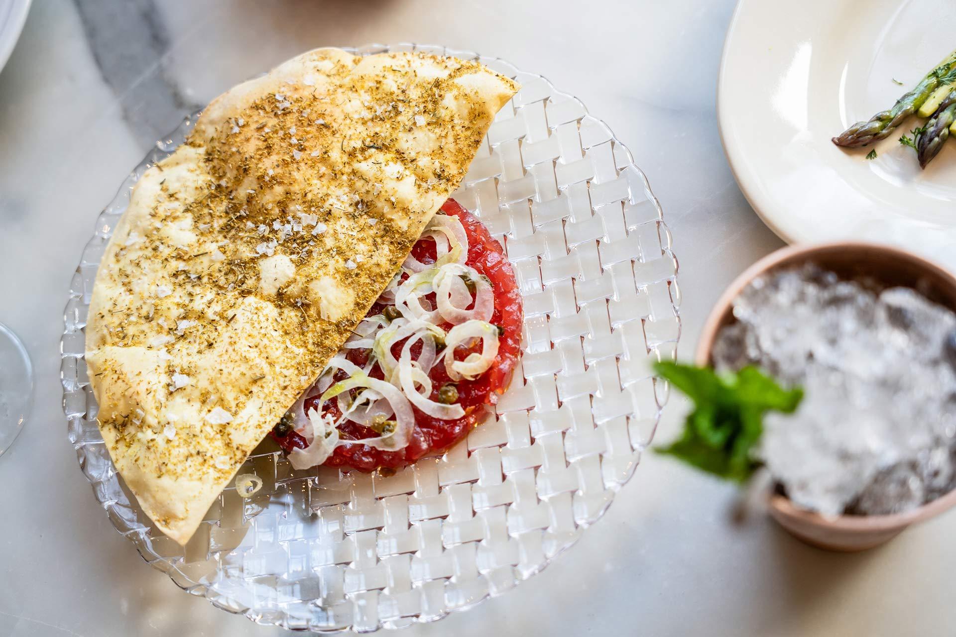 A clear glass plate with a half-folded flatbread topped with herbs and cheese, next to finely chopped raw meat and sliced onions. Other dishes and a drink with mint are partially visible nearby.