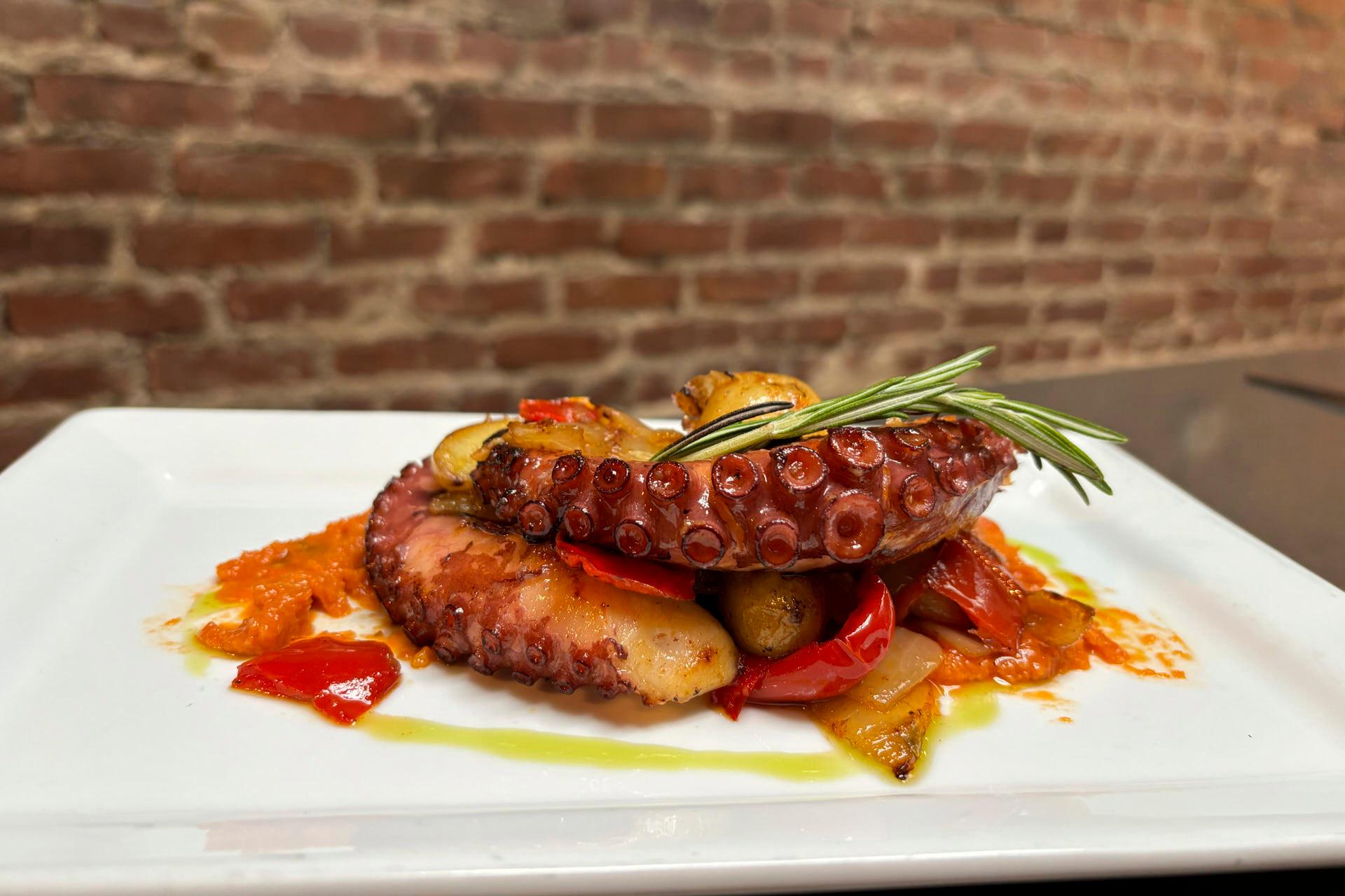 A dish featuring grilled octopus tentacles garnished with a sprig of rosemary, surrounded by roasted vegetables, including red peppers, on a white square plate. The background shows a brick wall.