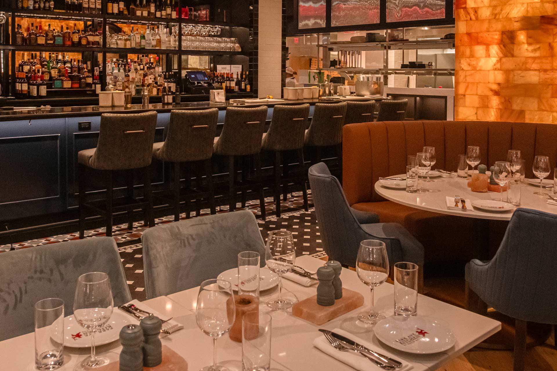 Cozy restaurant interior with elegantly set tables, cushioned chairs, and a bar lined with high-backed stools. Warm lighting highlights the decor, featuring neatly arranged glassware and tableware. Shelves behind the bar display an assortment of bottles.