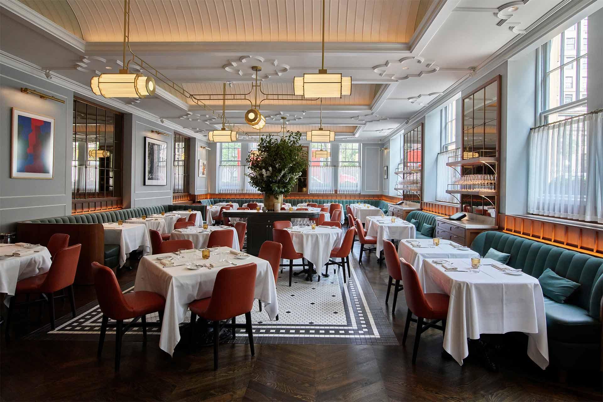 Elegant restaurant interior with white tablecloths, red chairs, and green banquette seating. Large central floral arrangement, modern lighting fixtures, wall art, and tall windows fill the space with natural light.