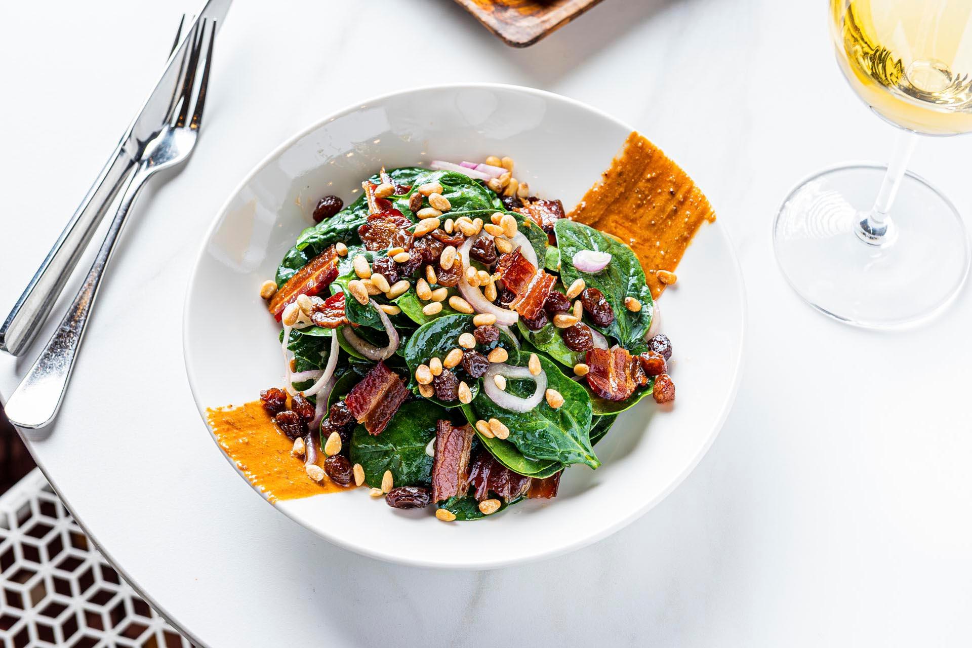 A bowl of spinach salad with crispy bacon, pine nuts, raisins, and sliced red onions. Garnished with cracker pieces. There's a glass of white wine to the right and cutlery on the left, on a white table.