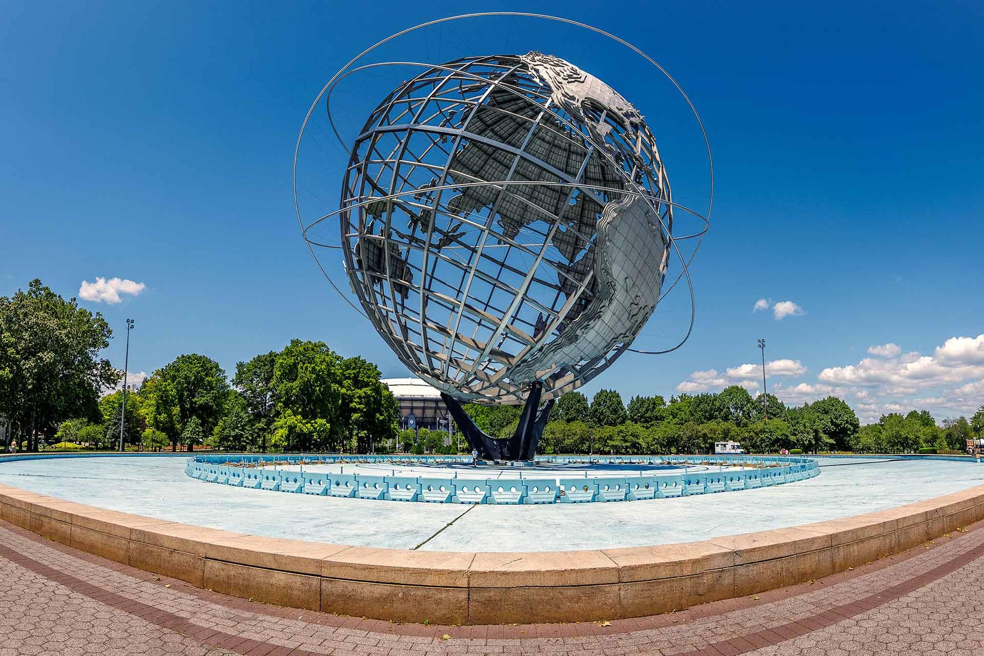 Large metal globe sculpture, called the Unisphere, stands in the center of a circular fountain surrounded by trees and a clear blue sky in a park setting.