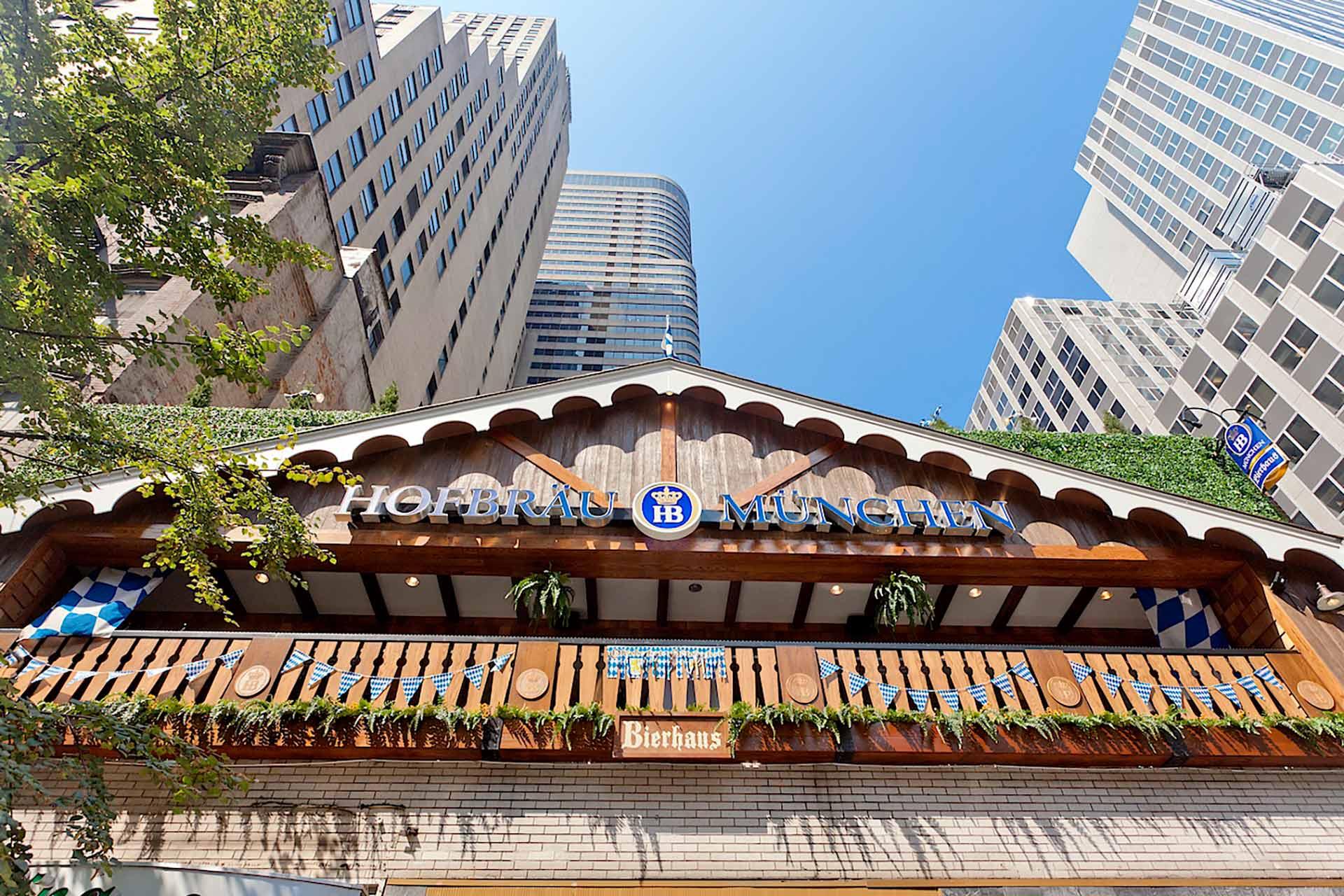 A wooden facade of Hofbräu München Bierhaus, decorated with blue and white flags and plants, set among tall modern buildings under a clear blue sky.