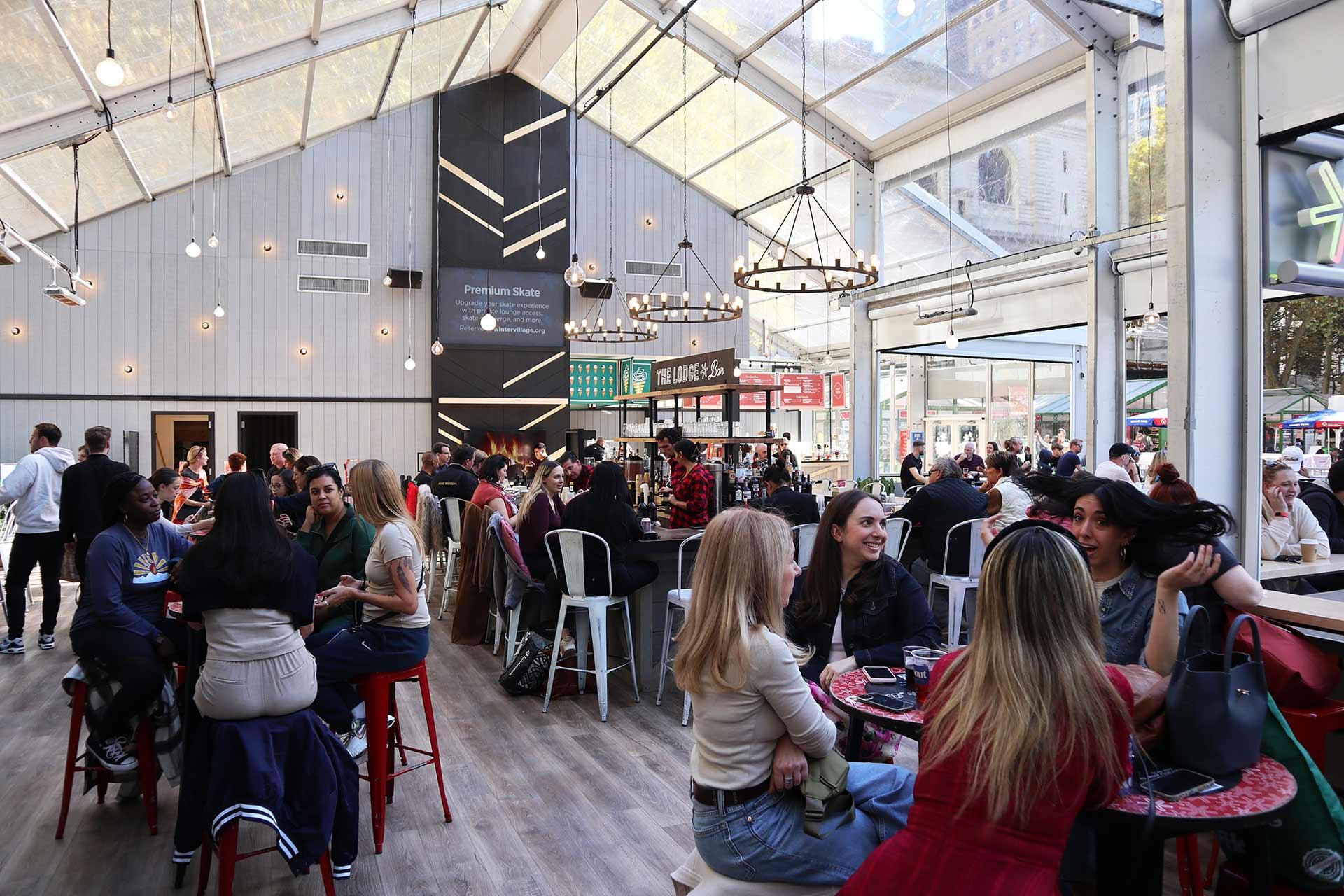 A bustling indoor food hall with large windows and high ceilings, filled with people sitting at tables and chatting; decorative lights hang from the ceiling and food stalls are visible in the background.