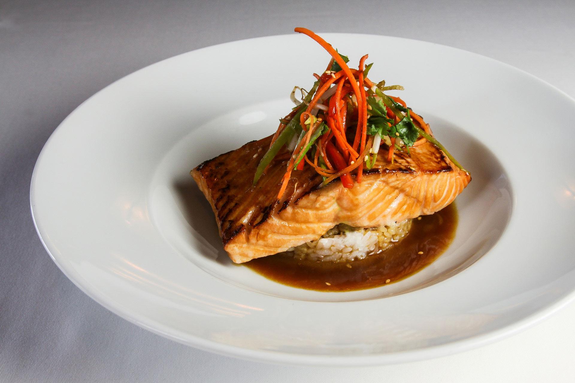 Grilled salmon fillet served on a bed of rice, topped with julienned vegetables, and surrounded by a brown sauce on a white plate.