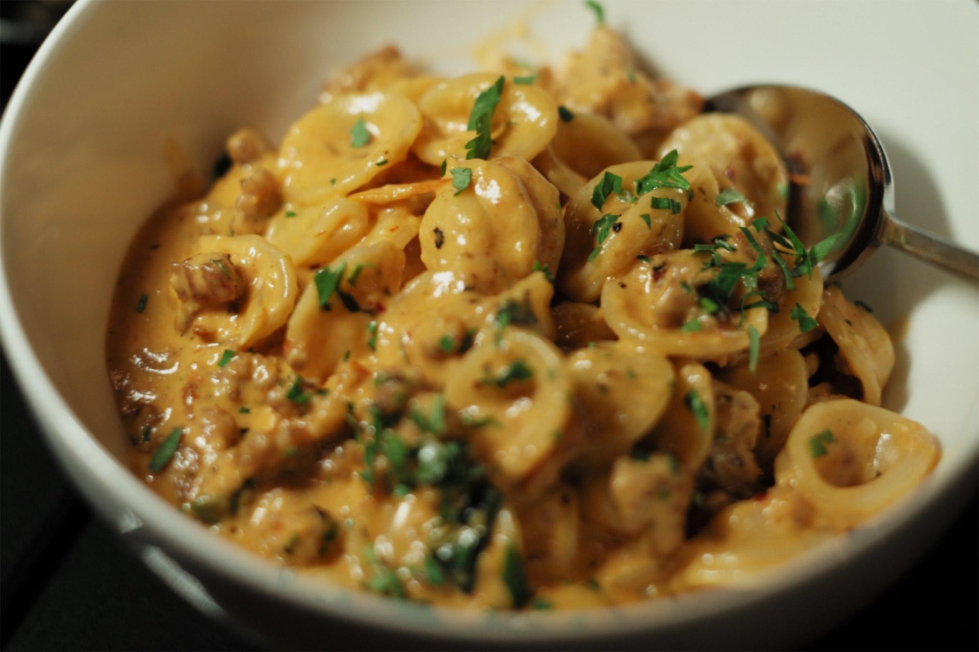 A bowl of creamy orecchiette pasta mixed with sausage and topped with chopped parsley, served with a spoon.