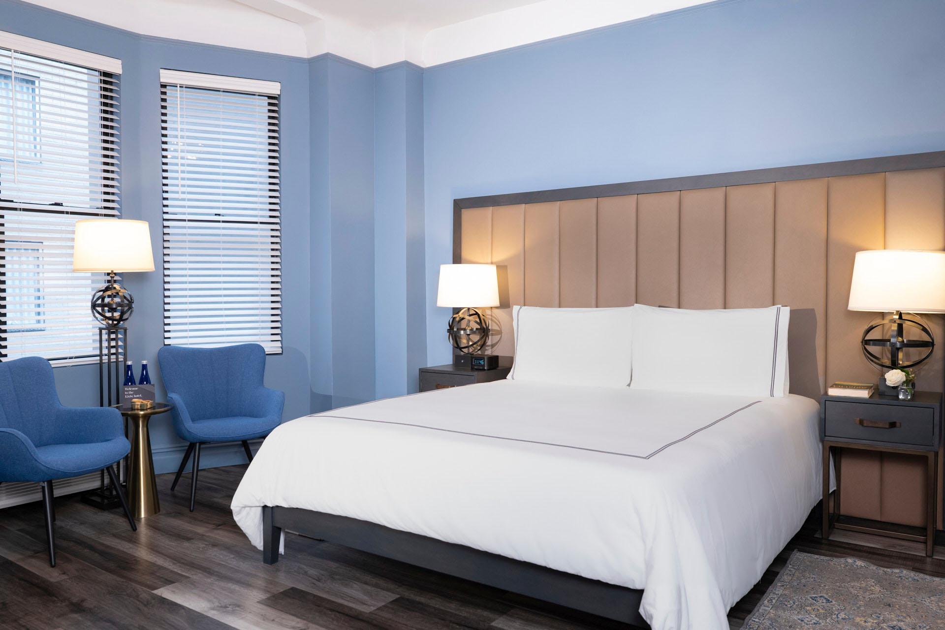 A modern hotel room with light blue walls, a large bed with white linens, two blue chairs by the window, two bedside tables with lamps, and wood flooring.