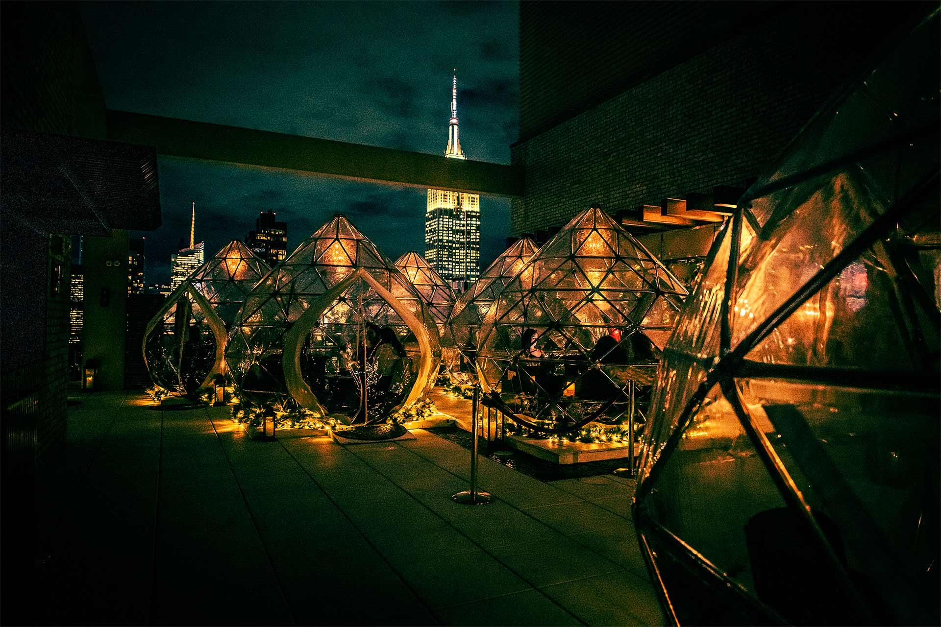 Rooftop at night with illuminated glass domes for dining, surrounded by string lights and plants, with skyscrapers and the Empire State Building glowing in the background.