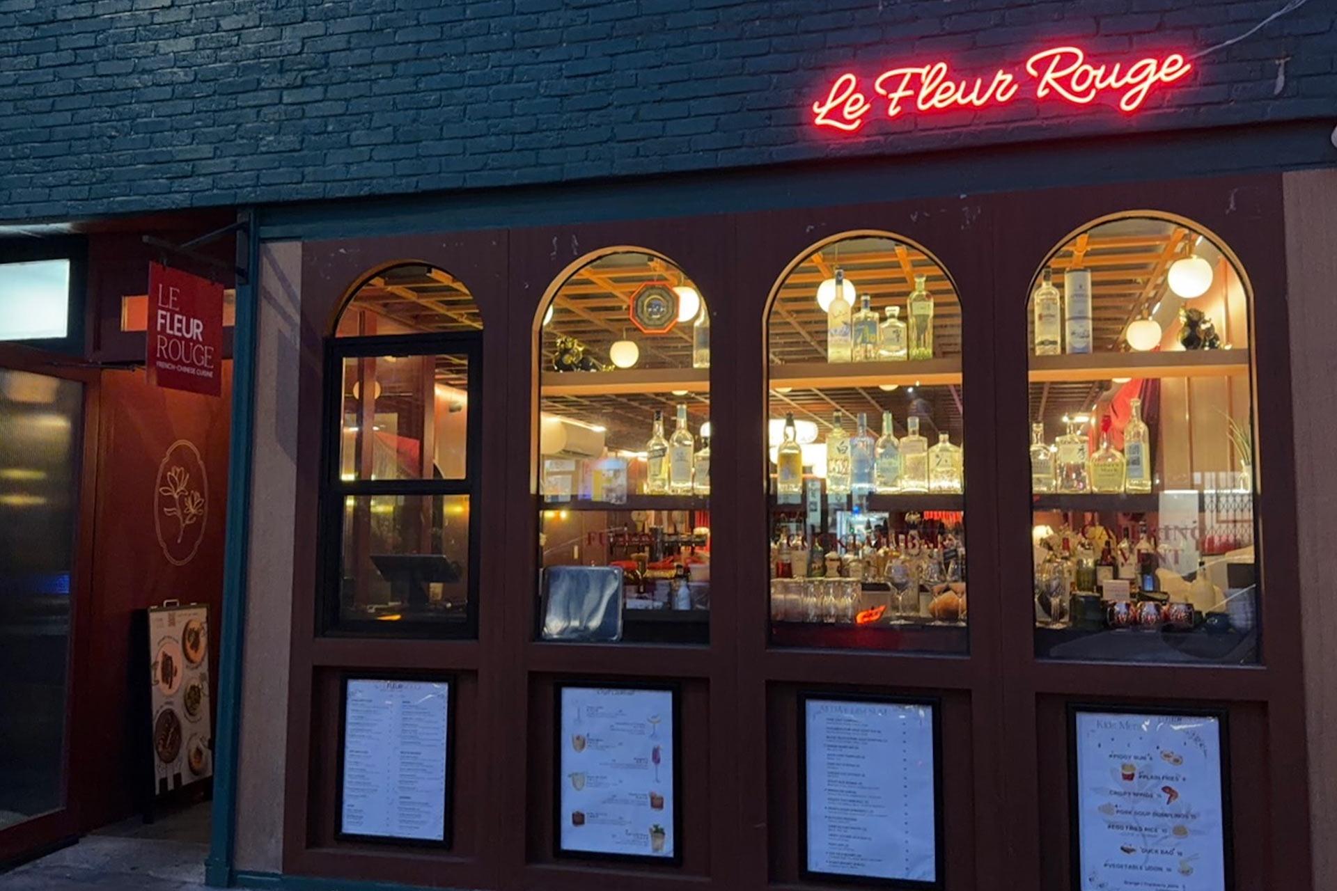 A restaurant called "Le Fleur Rouge" with a neon red sign, large arched windows displaying bottles on shelves, and printed menus posted on the exterior walls. Warm lights are visible inside.