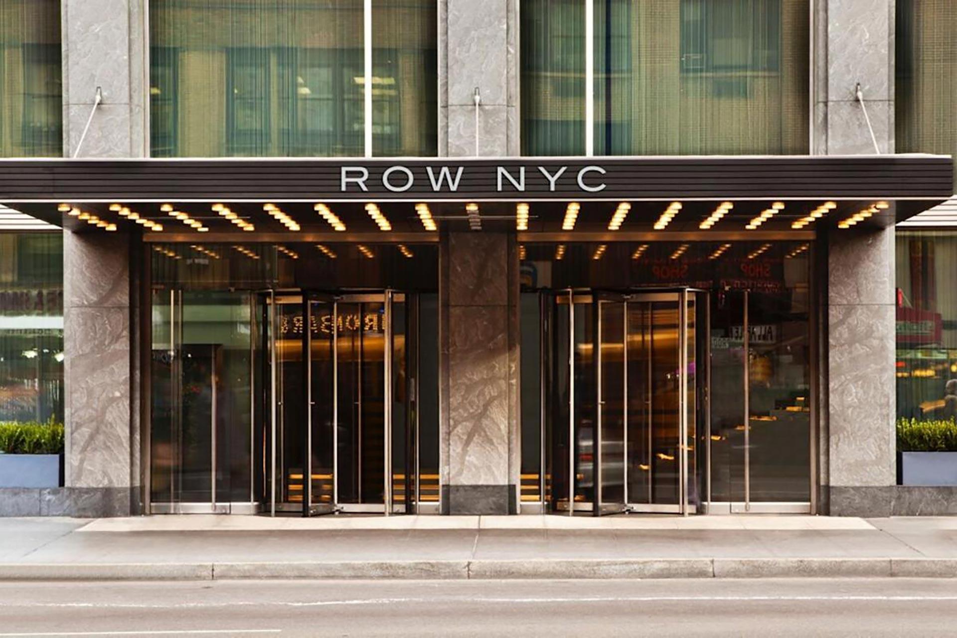 Entrance of the Row NYC hotel with a modern glass facade, metal accents, and a canopy with lights above the revolving doors. The hotel name “ROW NYC” is displayed prominently above the entrance.
