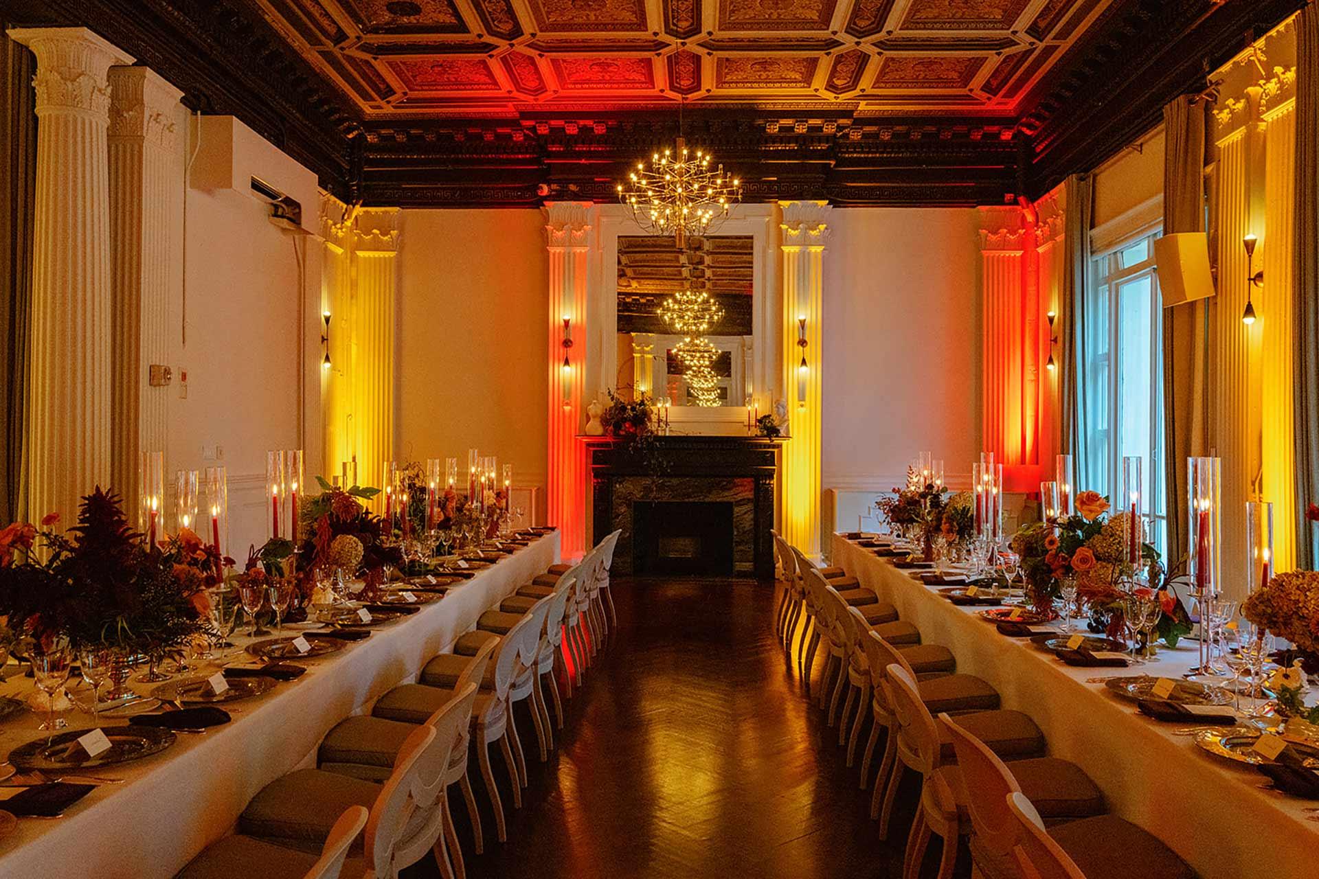 Elegant banquet hall with long dining tables set for an event, featuring floral centerpieces, tall candles, ornate chandeliers, high ceilings with intricate moldings, and warm yellow and orange lighting.