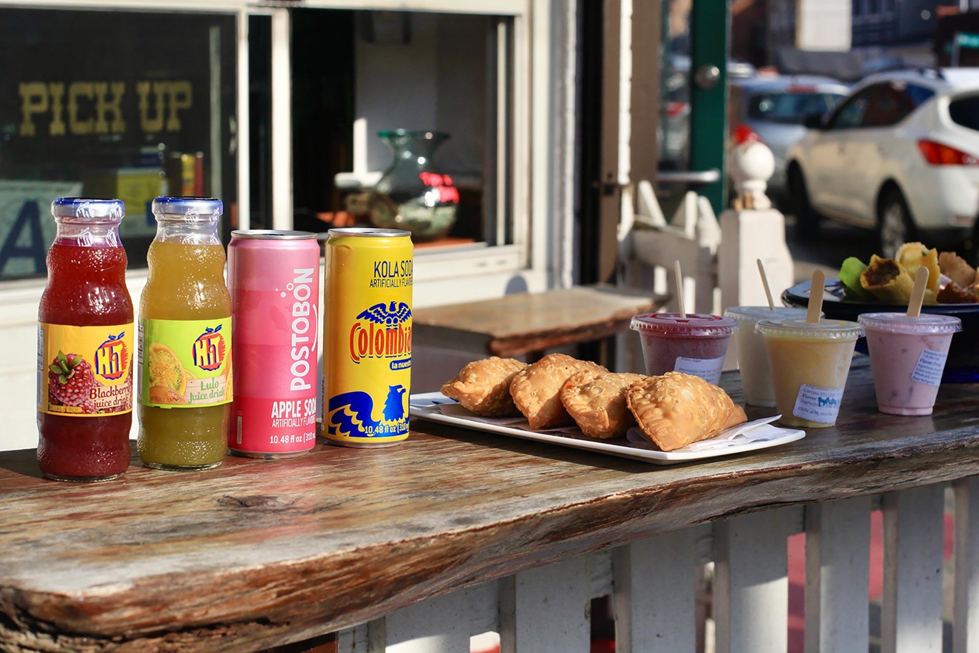 Four colorful bottled and canned drinks, a plate of empanadas, and several cups of smoothies and snacks are displayed on a rustic wooden table outside a shop, with cars and buildings visible in the background.