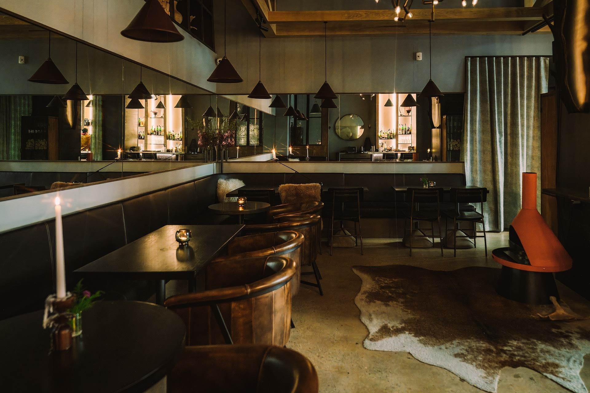 A dimly lit, modern lounge with leather chairs, dark tables, mirrors along the walls, pendant lights, a cowhide rug, and a small fireplace, creating a cozy, intimate atmosphere.