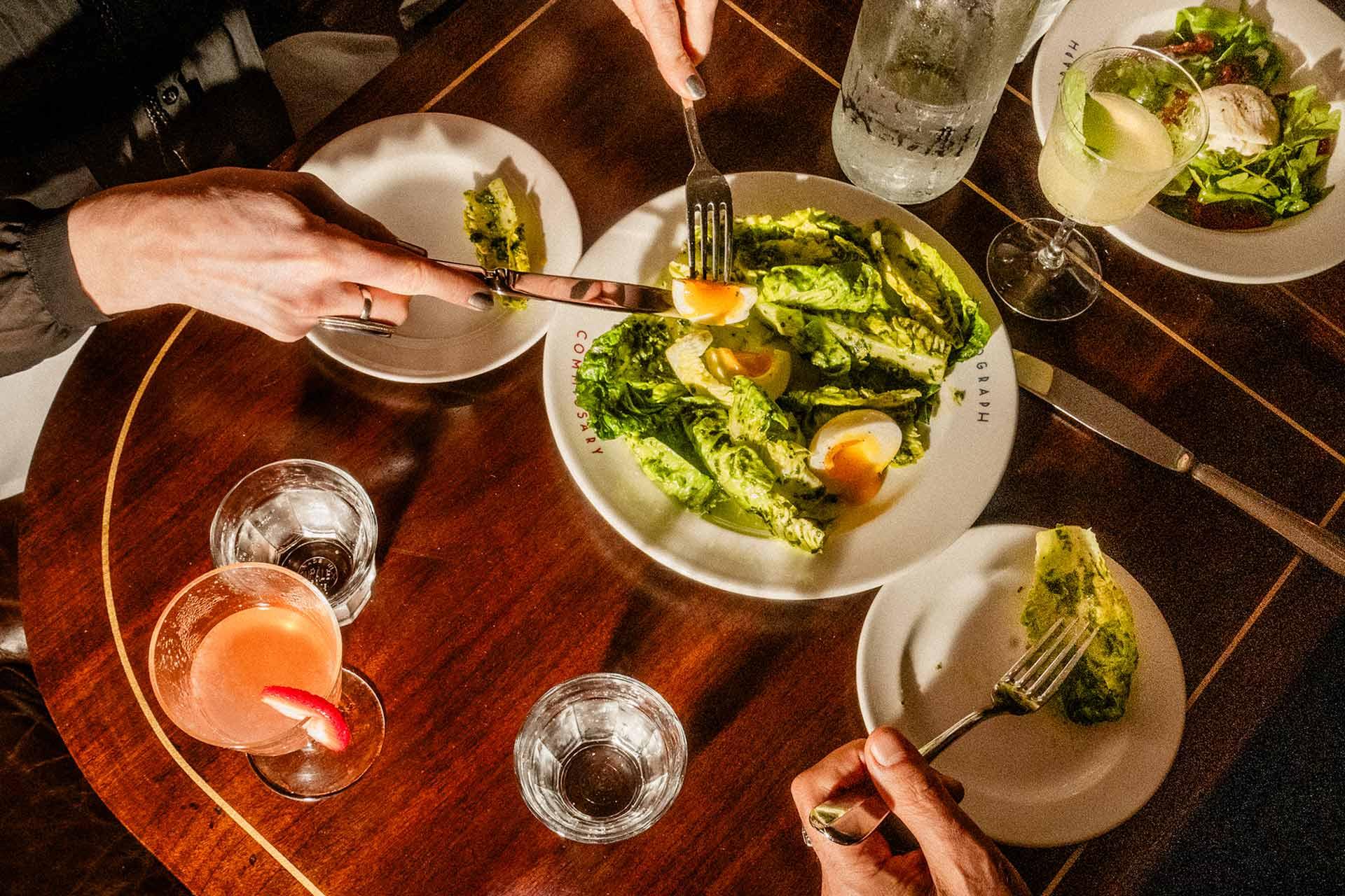 Two people sit at a wooden table, sharing a plate of salad with sliced eggs. Drinks, including a pink cocktail and a clear beverage with a lemon twist, are also on the table. Each person has a small plate and glass of water.