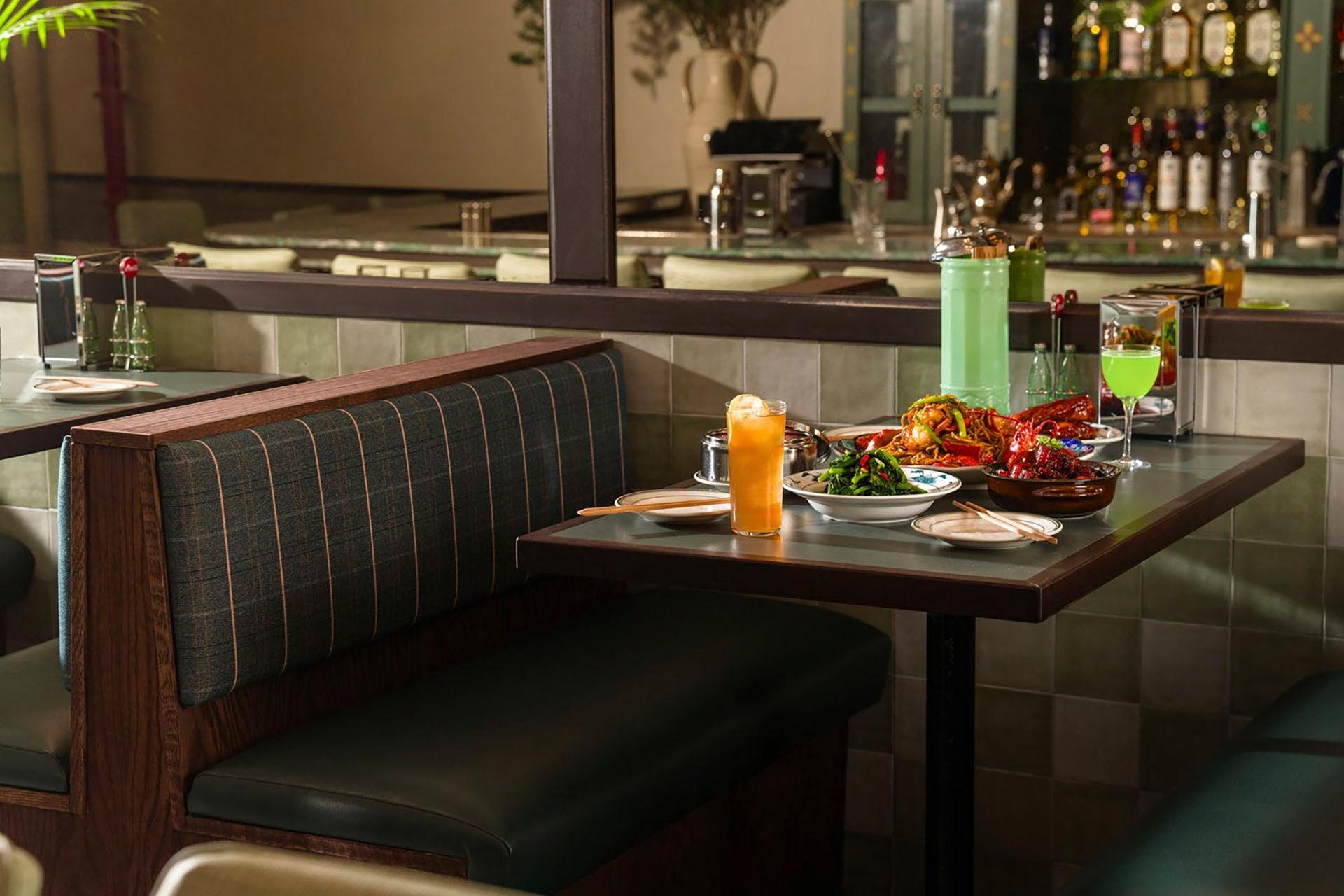 A cozy restaurant booth with green cushioned seats, featuring a table set with various dishes, including vibrant salads and meats. Two drinks are also present. The background shows a mirror and shelves with bottles.