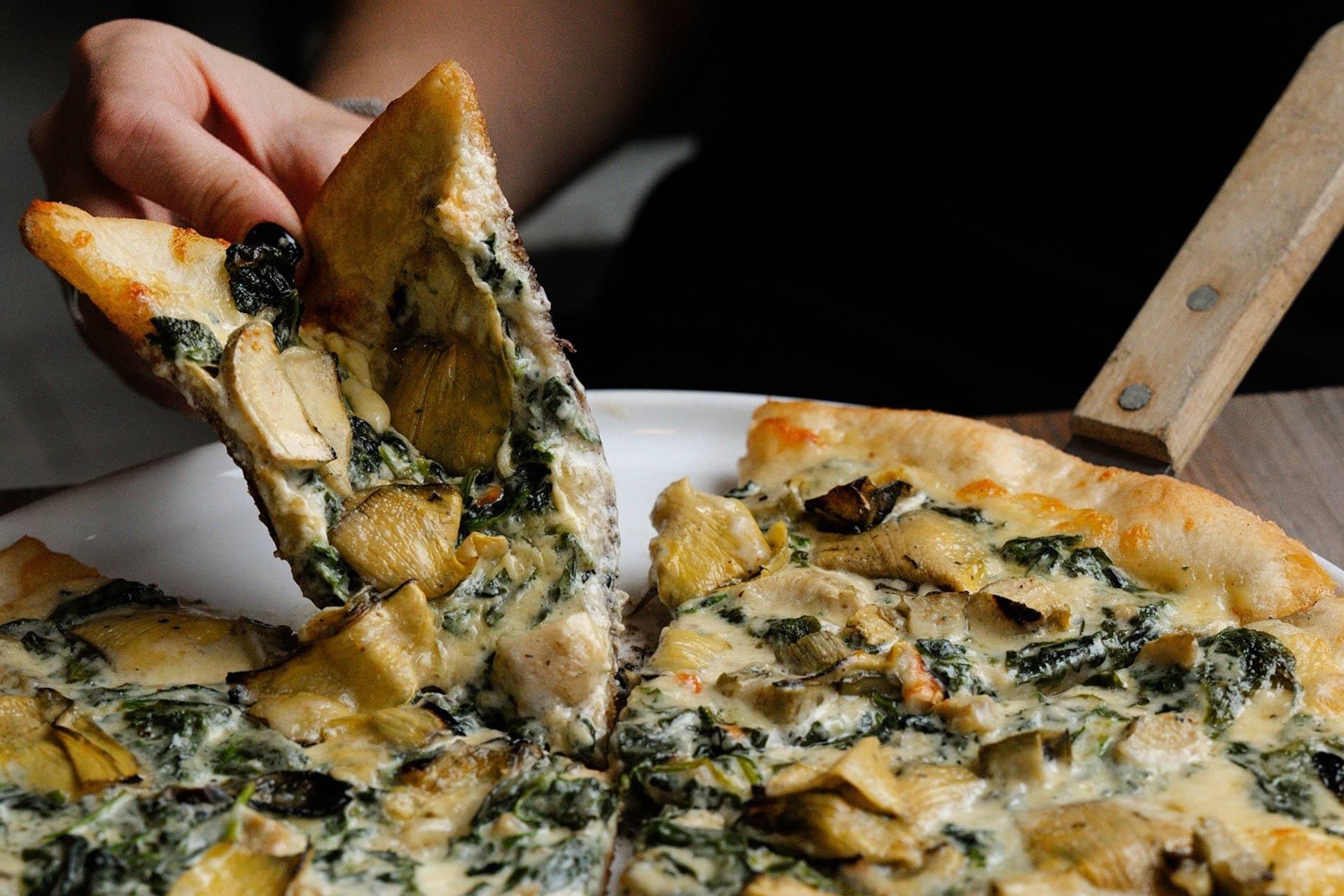 A hand lifts a slice of pizza topped with spinach, artichoke, mushrooms, and melted cheese from a whole pizza on a white plate.