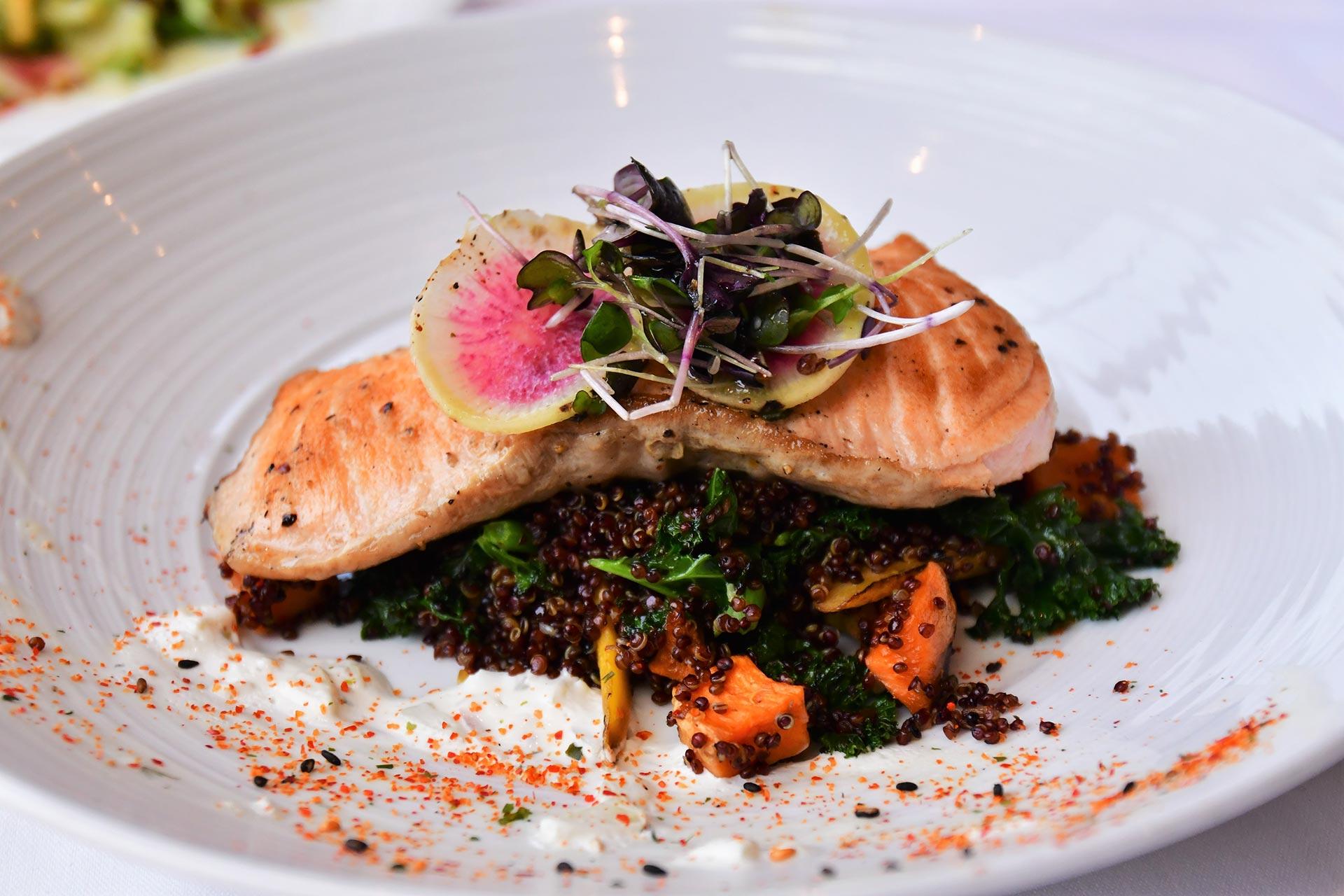 Grilled salmon fillet served atop a bed of quinoa and sautéed vegetables, garnished with microgreens and a thin radish slice on a white plate, with a sprinkle of seasoning around the edges.