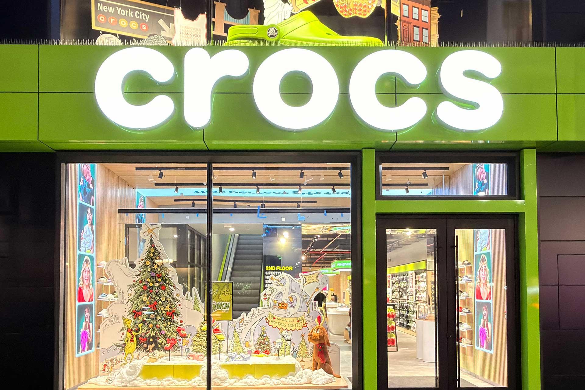 A brightly lit Crocs store with a large white logo above the entrance, festive holiday decorations, a Christmas tree, and colorful displays visible inside through the windows.