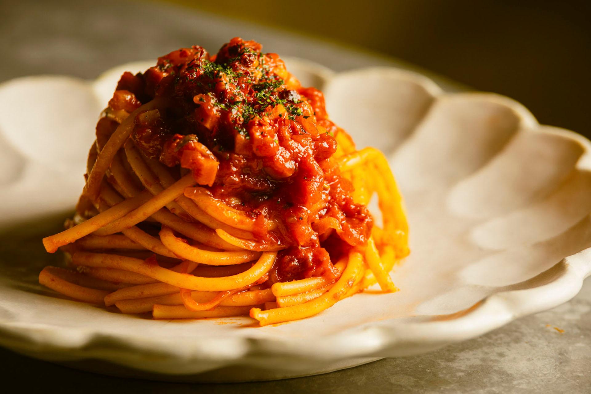 A plate of spaghetti topped with a rich, chunky tomato sauce, garnished with herbs, served on a white, scalloped-edge plate. The warm lighting enhances the dish's appetizing appearance.