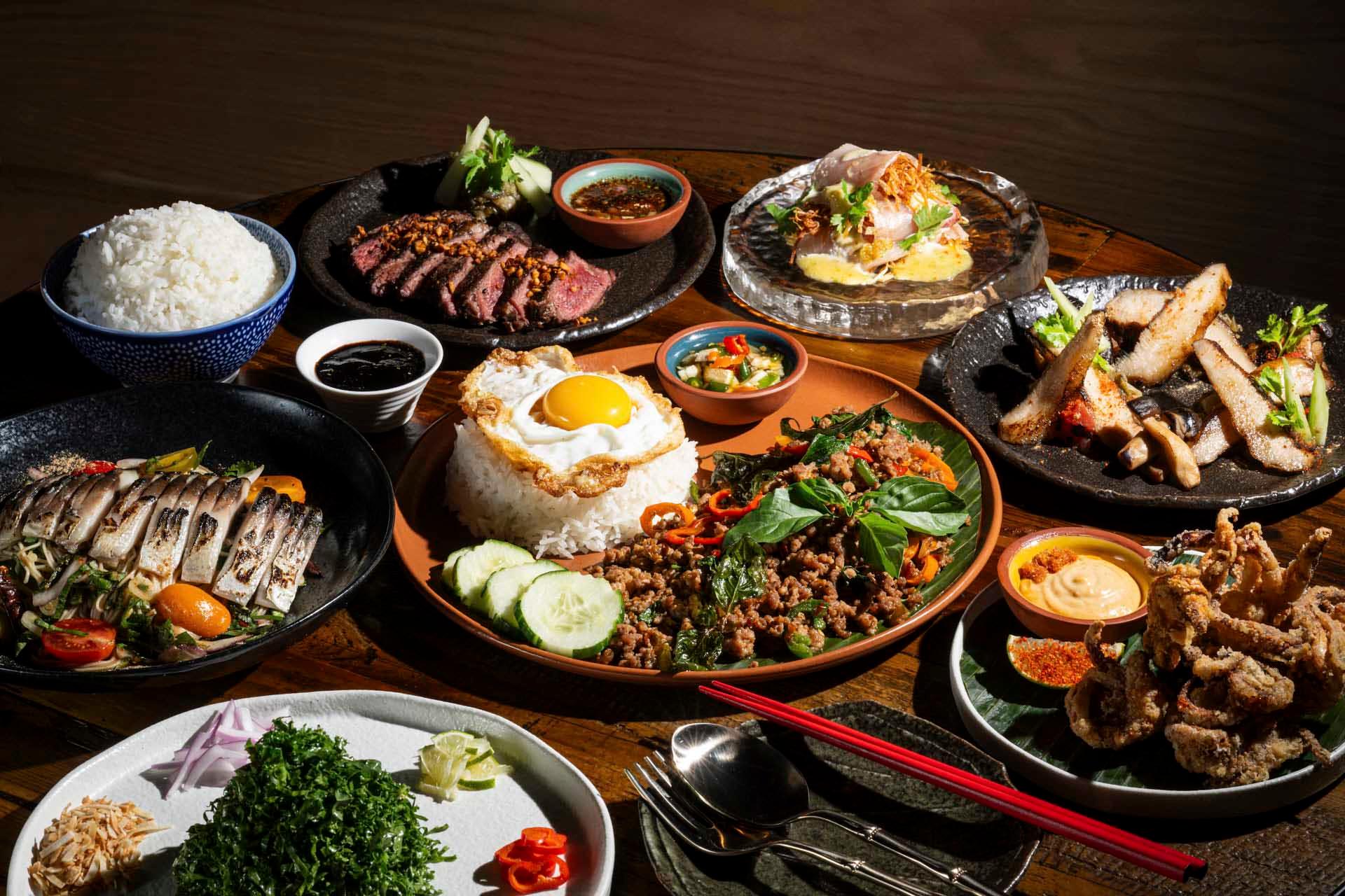 A variety of Asian dishes are arranged on a wooden table. There's rice with a fried egg, grilled meat, leafy greens, fried fish, and dipping sauces. Red chopsticks and a fork are placed nearby.