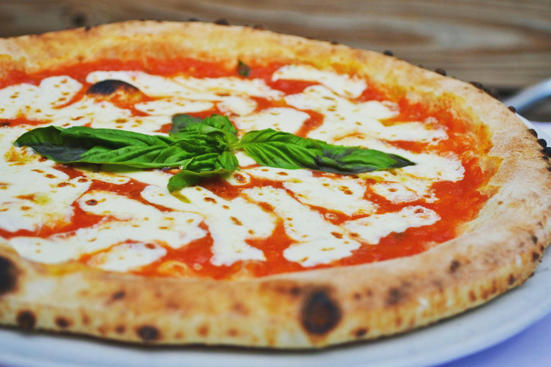 A freshly baked margherita pizza with melted mozzarella cheese, vibrant red tomato sauce, and fresh basil leaves on top. The crust is golden-brown and slightly charred, served on a white plate.