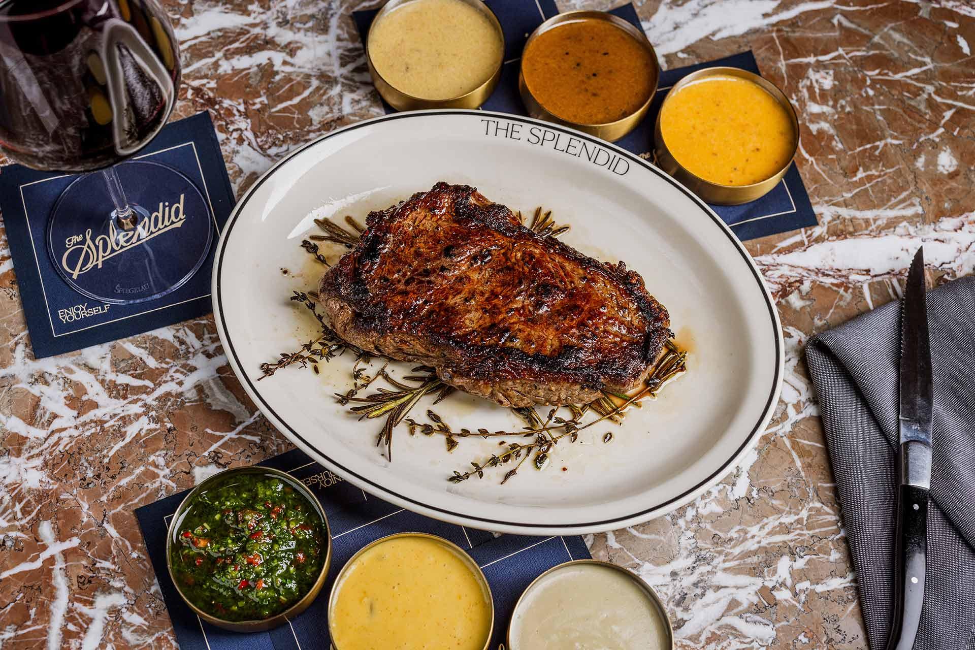 A grilled steak served on a white plate garnished with herbs, surrounded by five different sauces in small bowls on a marble table. A knife, napkin, and drink are nearby. The plate is labeled “The Splendid.”.
