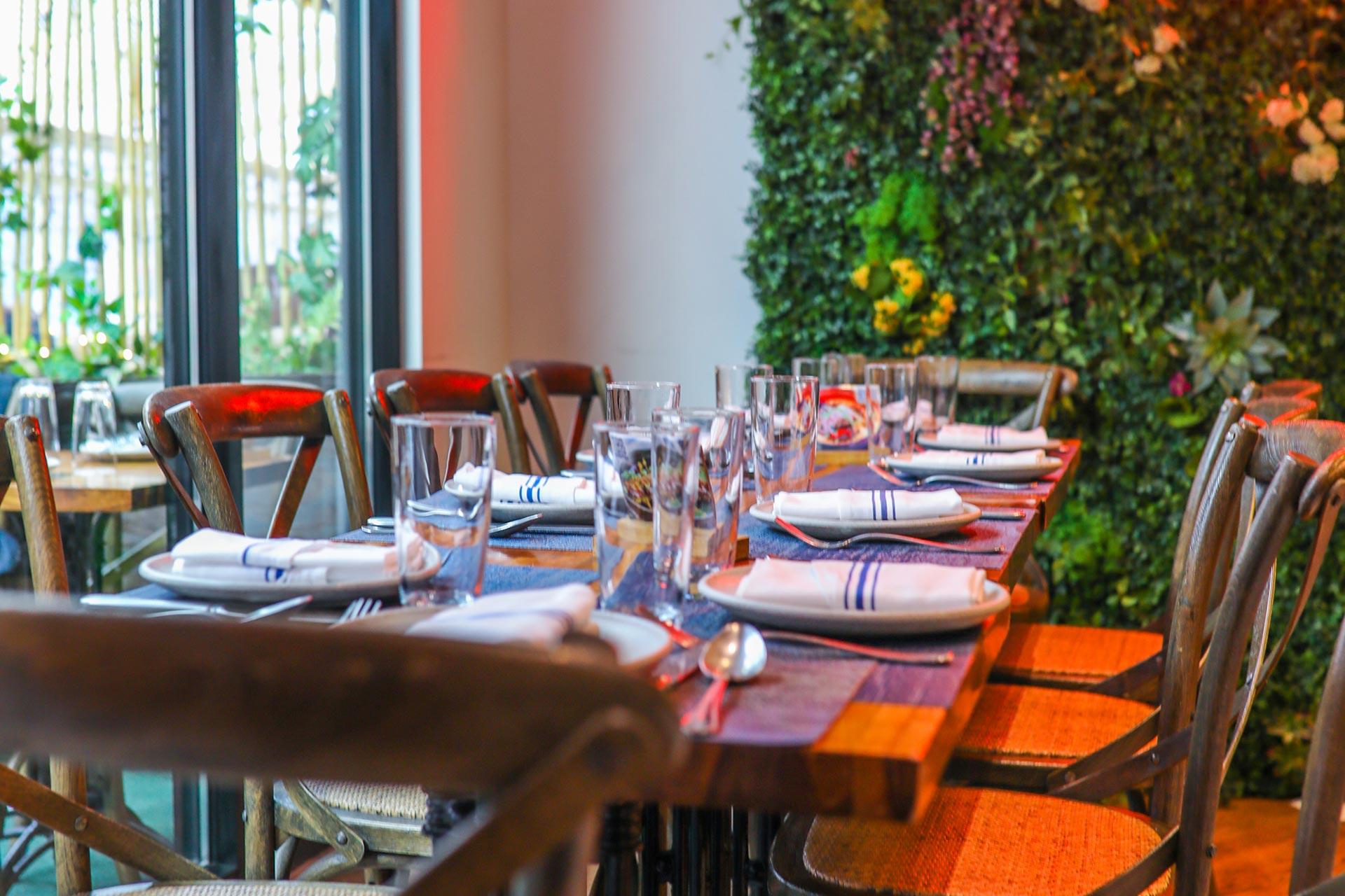 A wooden dining table set with plates, napkins, glasses, and cutlery, surrounded by wooden chairs in a restaurant with a green wall decorated with plants and flowers in the background.