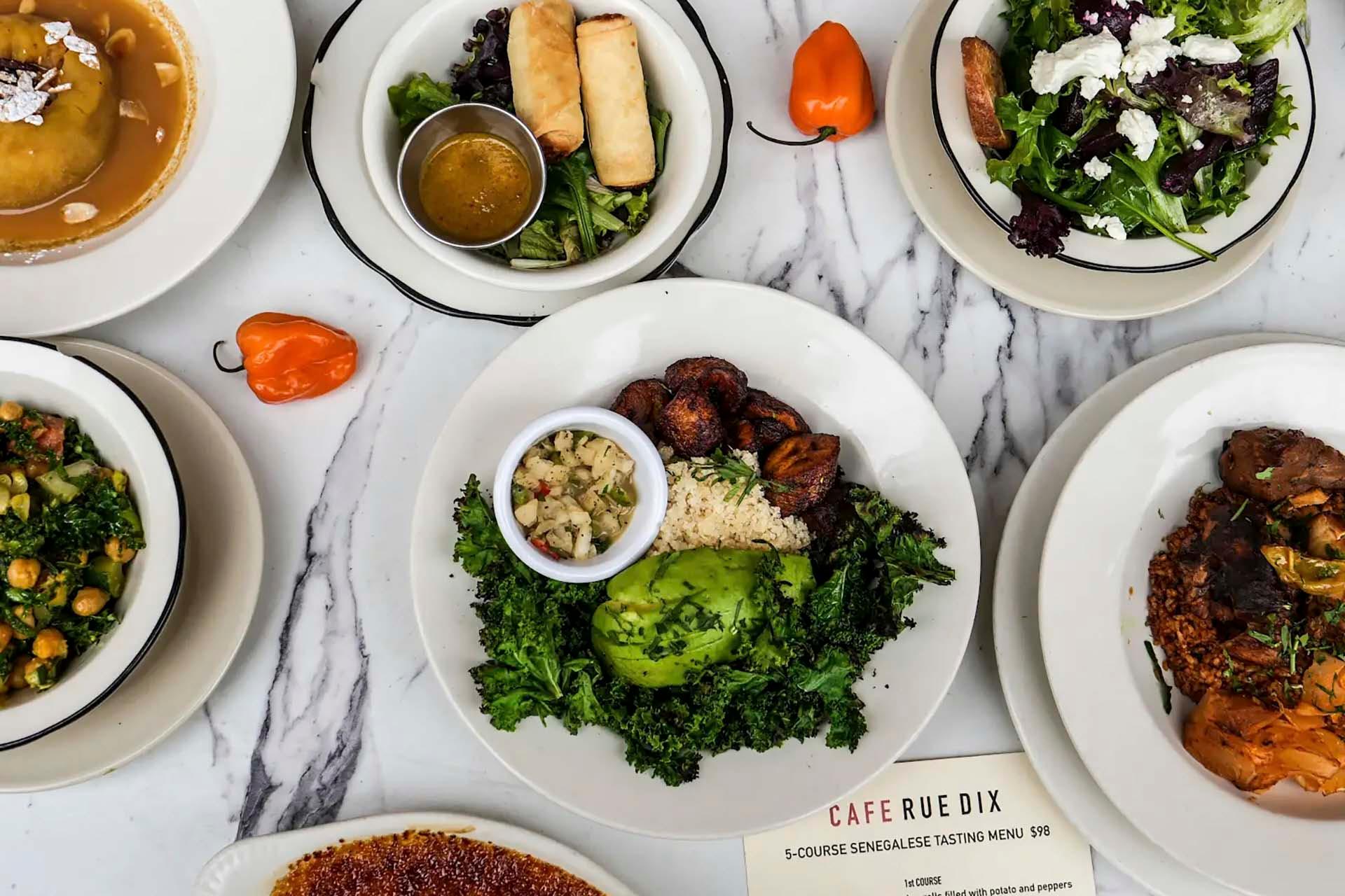 An array of dishes on a marble table, including plates of salad, plantains with avocado and kale, spring rolls, and a bowl of soup. A menu is visible at the bottom.