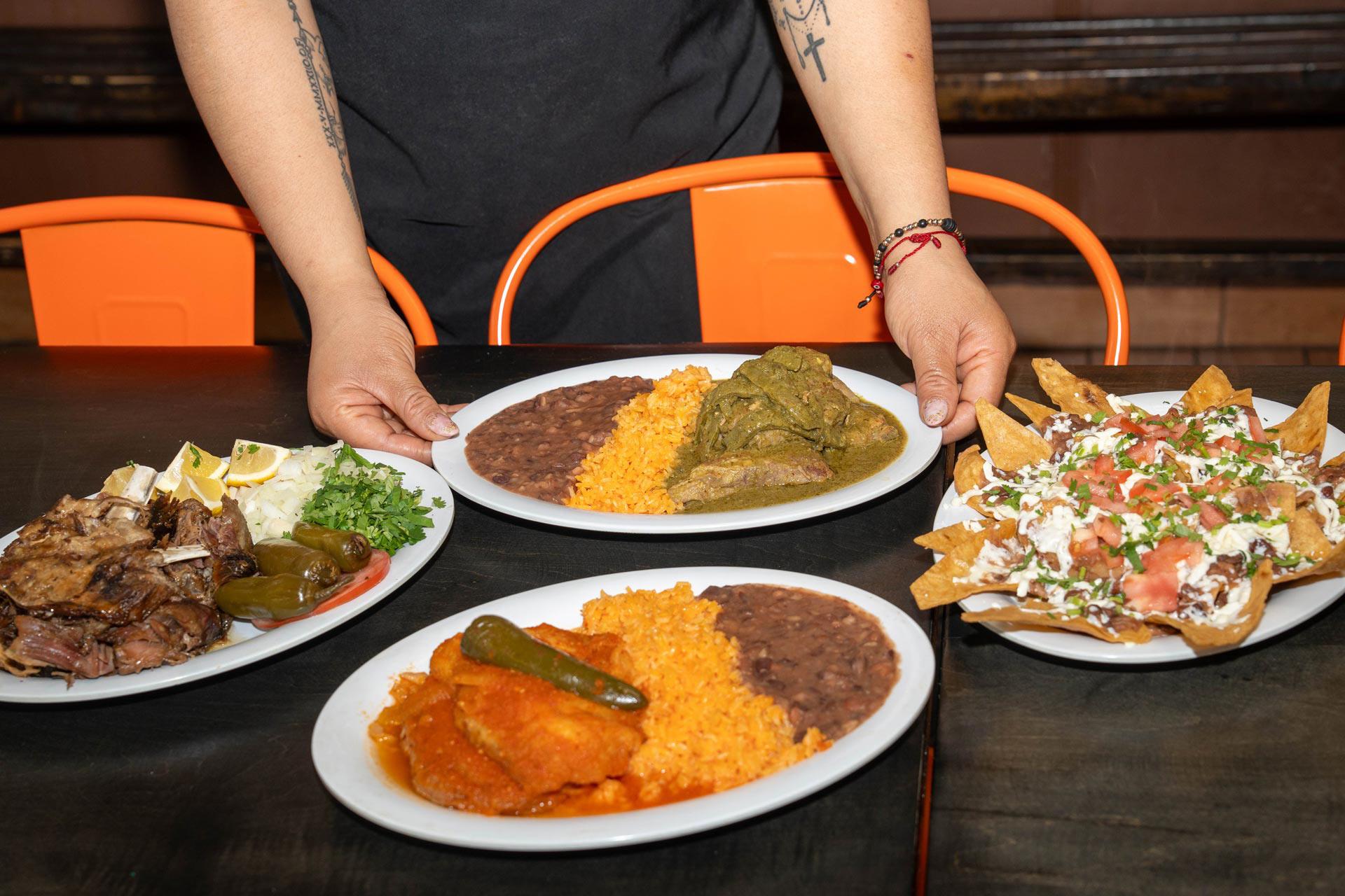 A person with tattoos on their arms sets down a plate of rice, beans, and green sauce meat among other dishes, including nachos on a dark table with orange chairs.