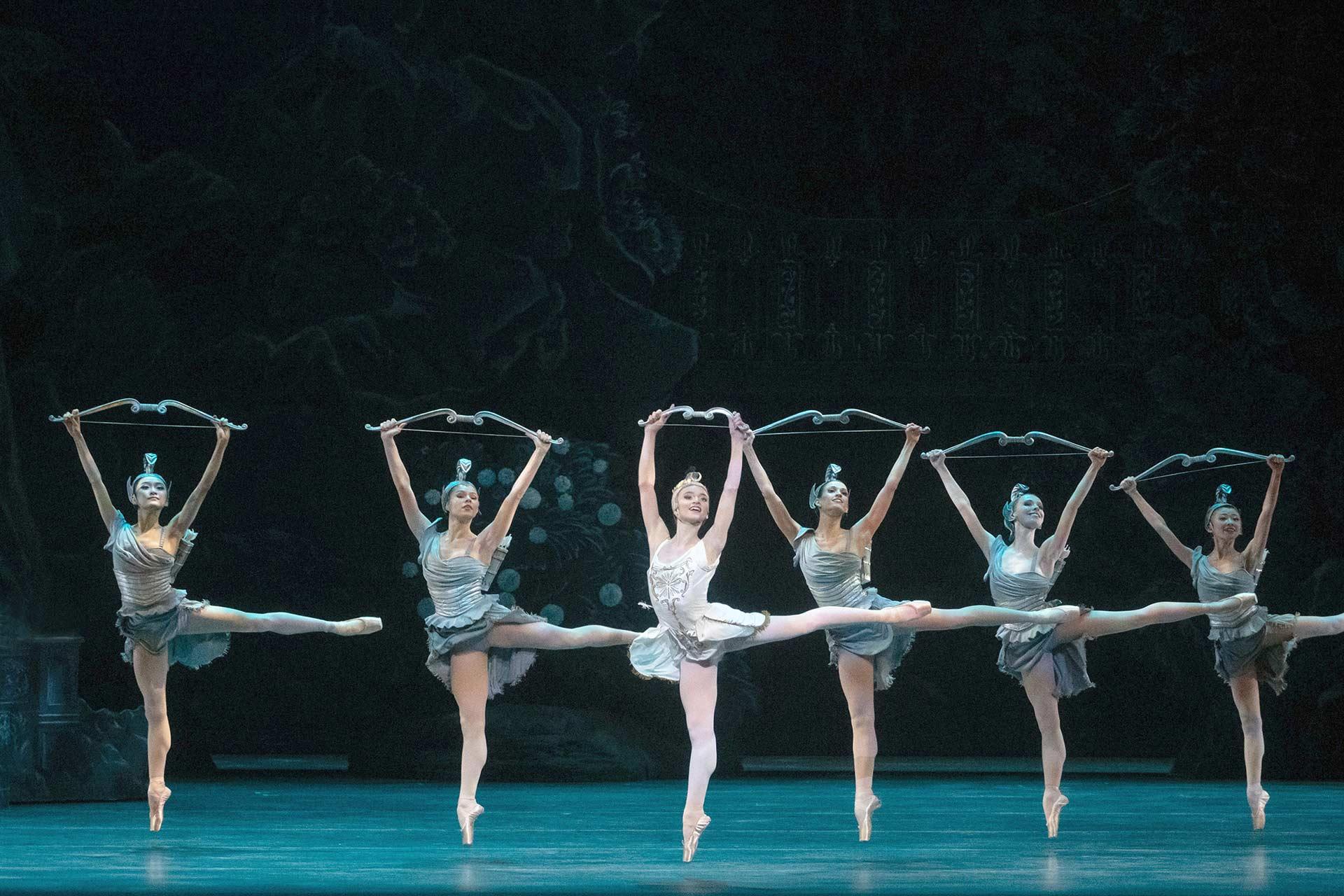 Six ballerinas in tutus perform on stage, each holding a garland above their heads and extending one leg high to the side in a ballet pose, with a dark, ornate backdrop behind them.