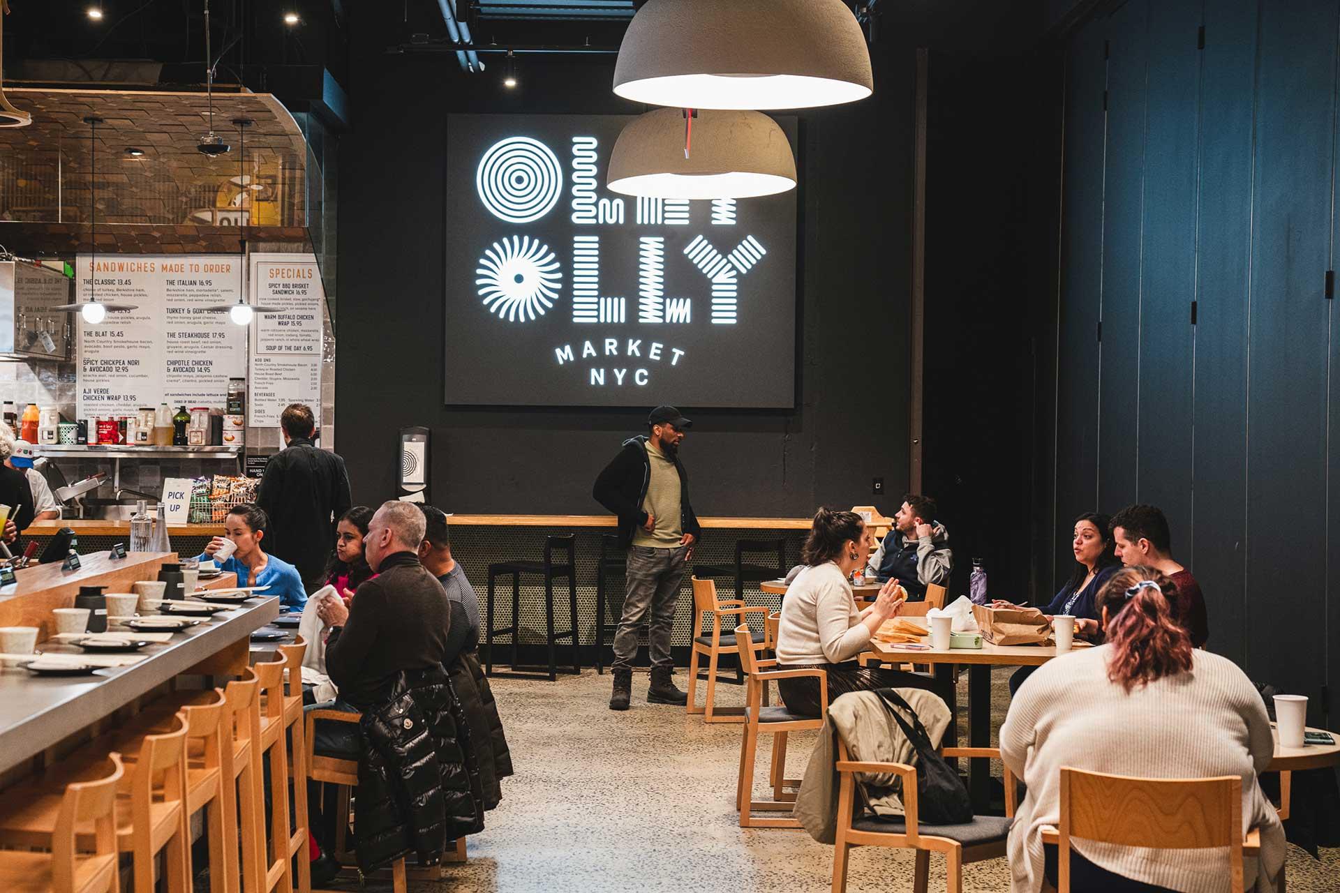 A modern indoor food market with people dining at tables and a bar, featuring a large illuminated sign reading “Ollie Market NYC” on the back wall. One person stands near the sign while others are seated and eating.