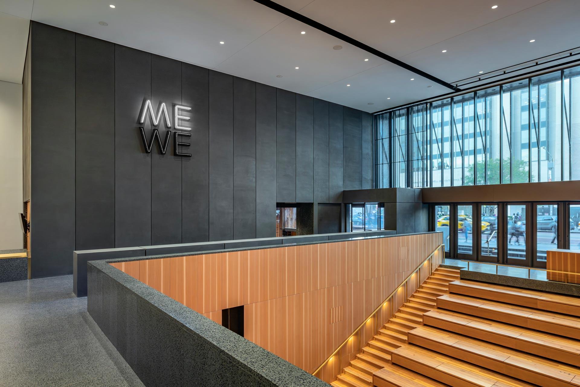 Modern lobby with large windows, wooden bleacher-style seating, dark walls, and a glowing sign reading "ME WE." Yellow taxis and city buildings are visible outside through the glass entrance.