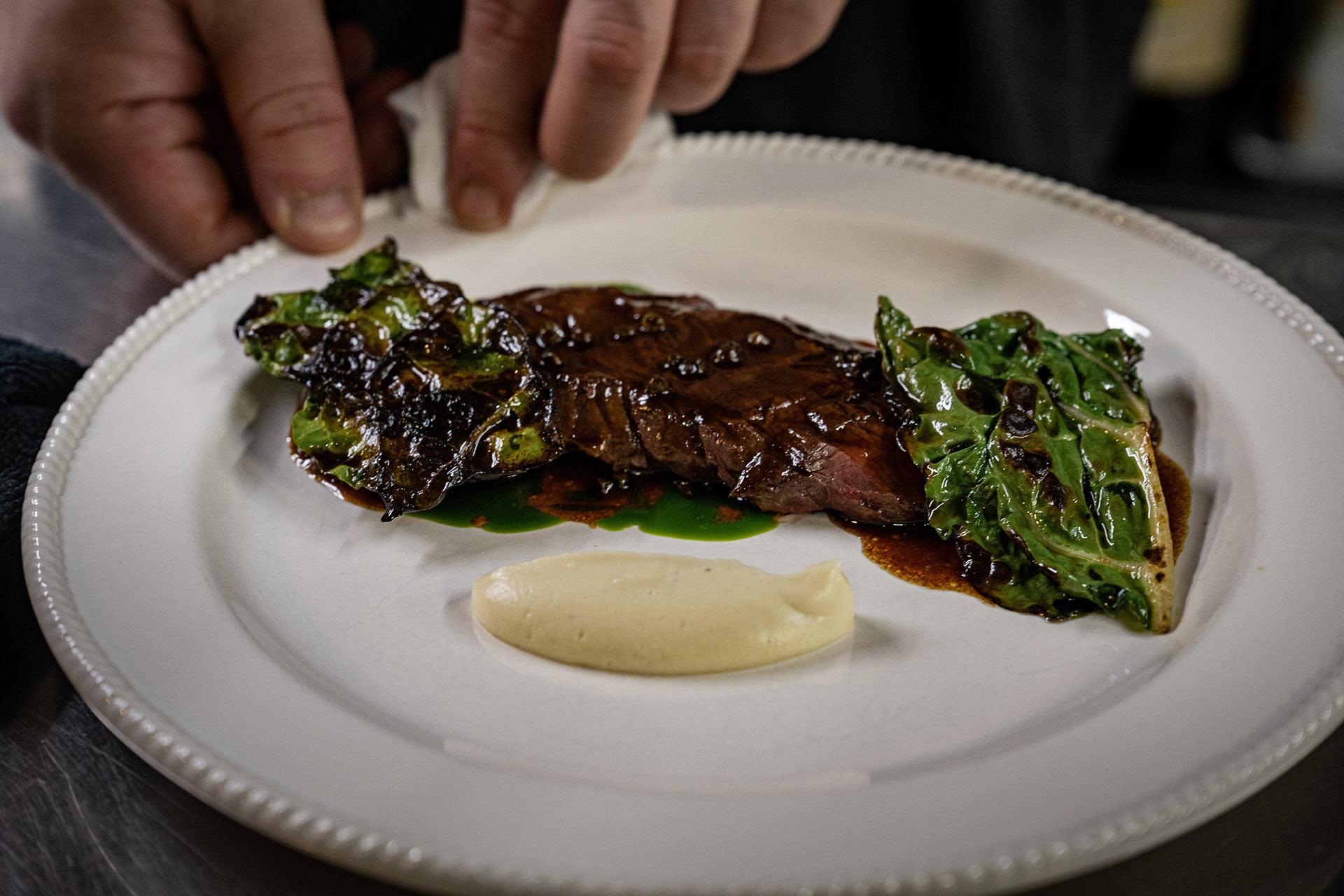 A chef’s hands plate grilled leafy greens and a glazed steak beside a swirl of creamy mashed potatoes on a white dish.