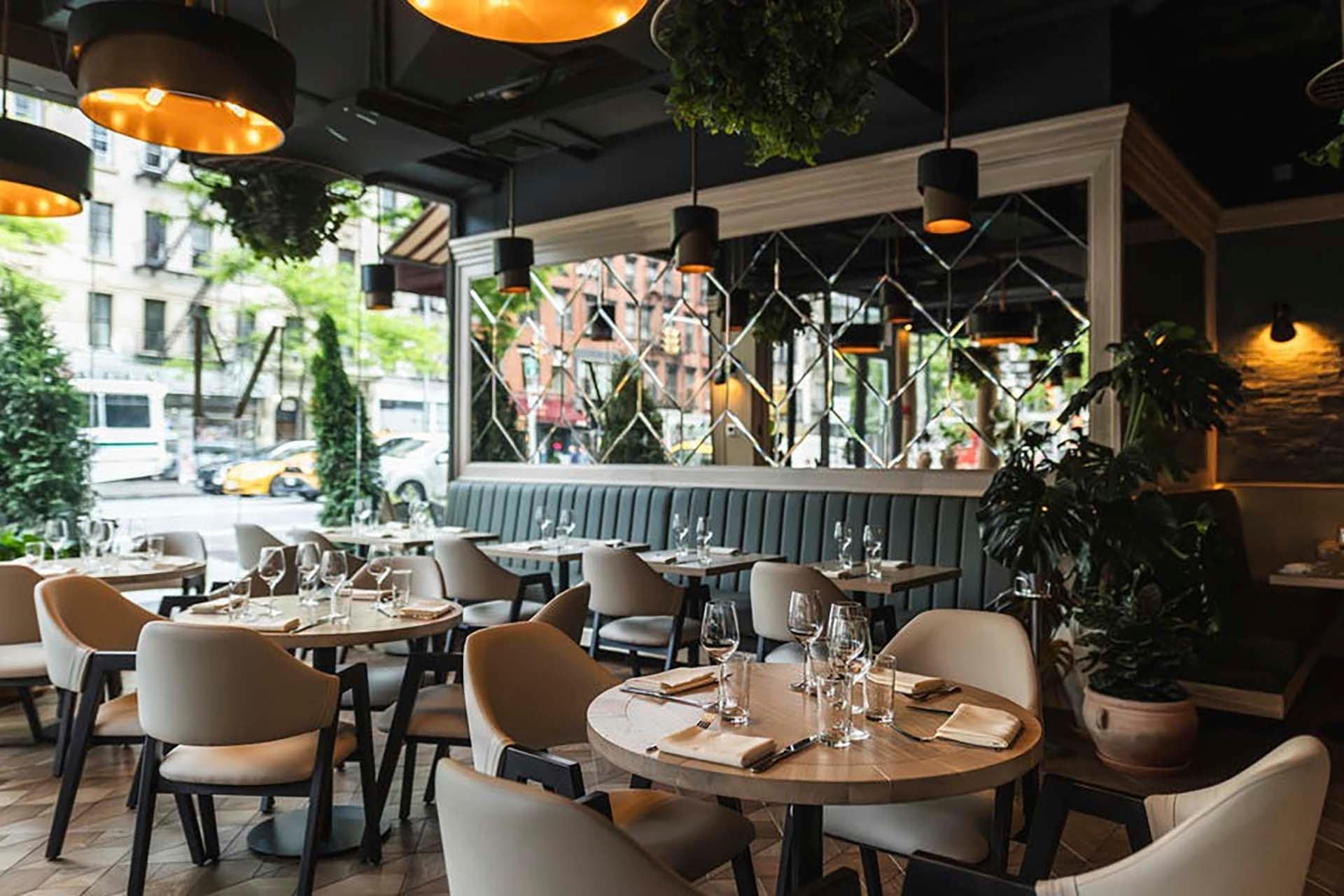 Cozy restaurant interior with modern decor. Beige chairs and round wooden tables are set with wine glasses and napkins. Large windows, hanging lights, and green plants add to the warm ambiance. Outside, city streets with trees and buildings are visible.