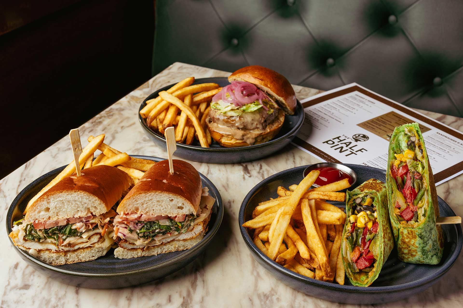 Three plates of food on a marble table: a sandwich with fries, a burger with fries, and a wrap with fries. A menu is in the background on a green cushioned seat.