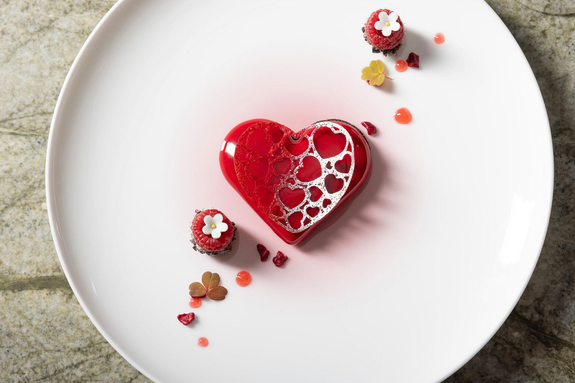 A heart-shaped red dessert with a decorative lace pattern sits on a white plate, accompanied by small round pastries, flower decorations, and heart-shaped sauce drops.