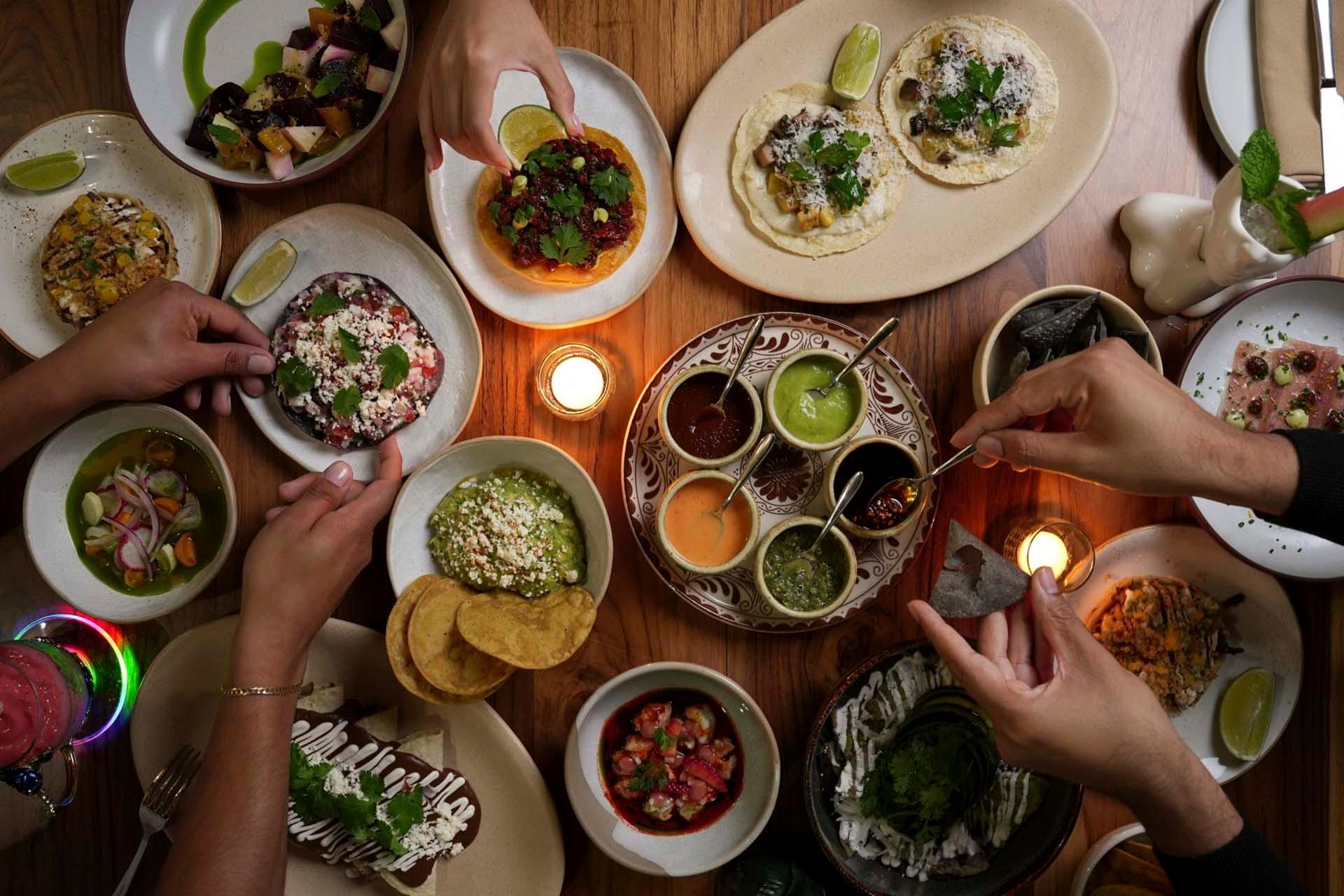 A tabletop filled with various Mexican dishes, including tacos, ceviche, and sauces. Several hands reach in to serve or sample the colorful food. Candles provide a warm ambiance, and drinks are visible at the edges of the table.