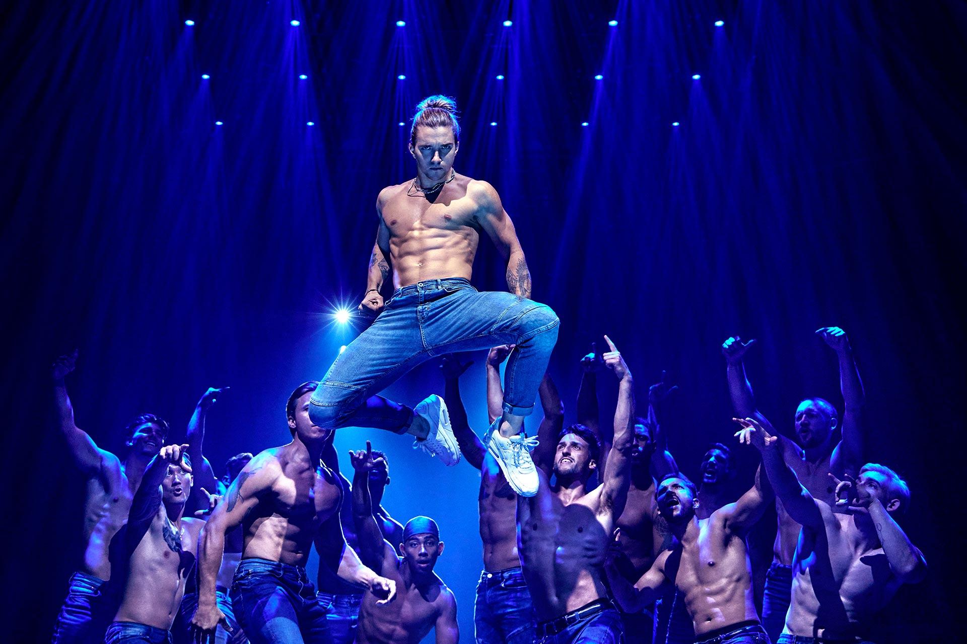 A shirtless man in jeans and sneakers is suspended mid-air under blue stage lights, surrounded by shirtless dancers reaching up toward him, creating a dramatic and energetic scene.