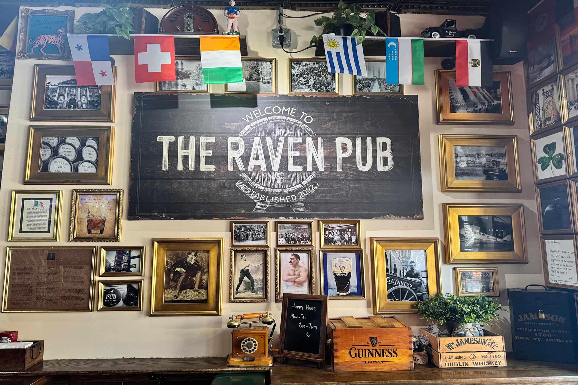 Wall of The Raven Pub decorated with framed photos, memorabilia, shelves, and small flags. A large sign reads "The Raven Pub." Guinness and Jameson boxes are on a counter beneath the sign.
