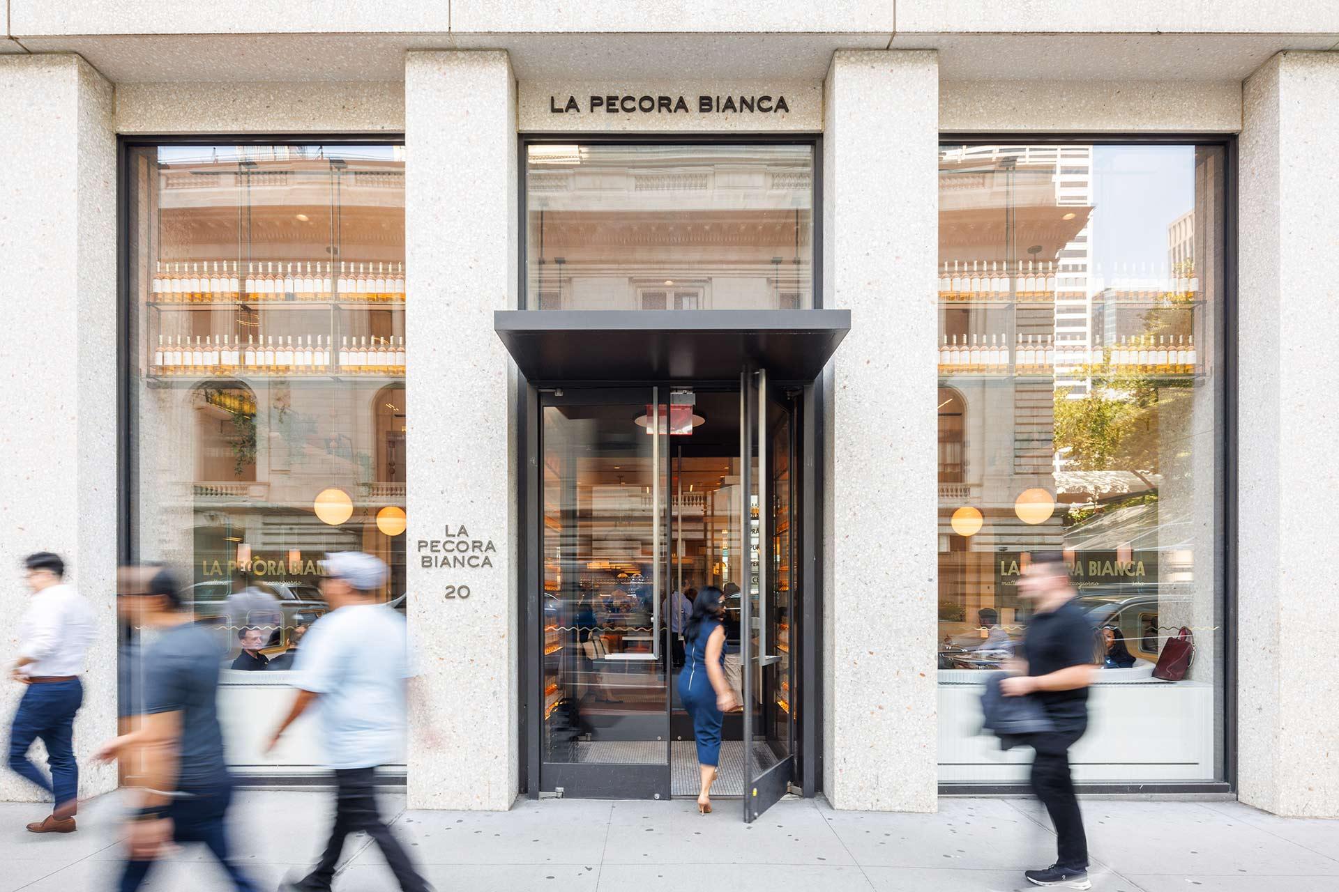 People walk past the entrance of La Pecora Bianca restaurant, which features large windows and a modern facade. One person is entering through the glass door, and the interior is visible with diners inside.