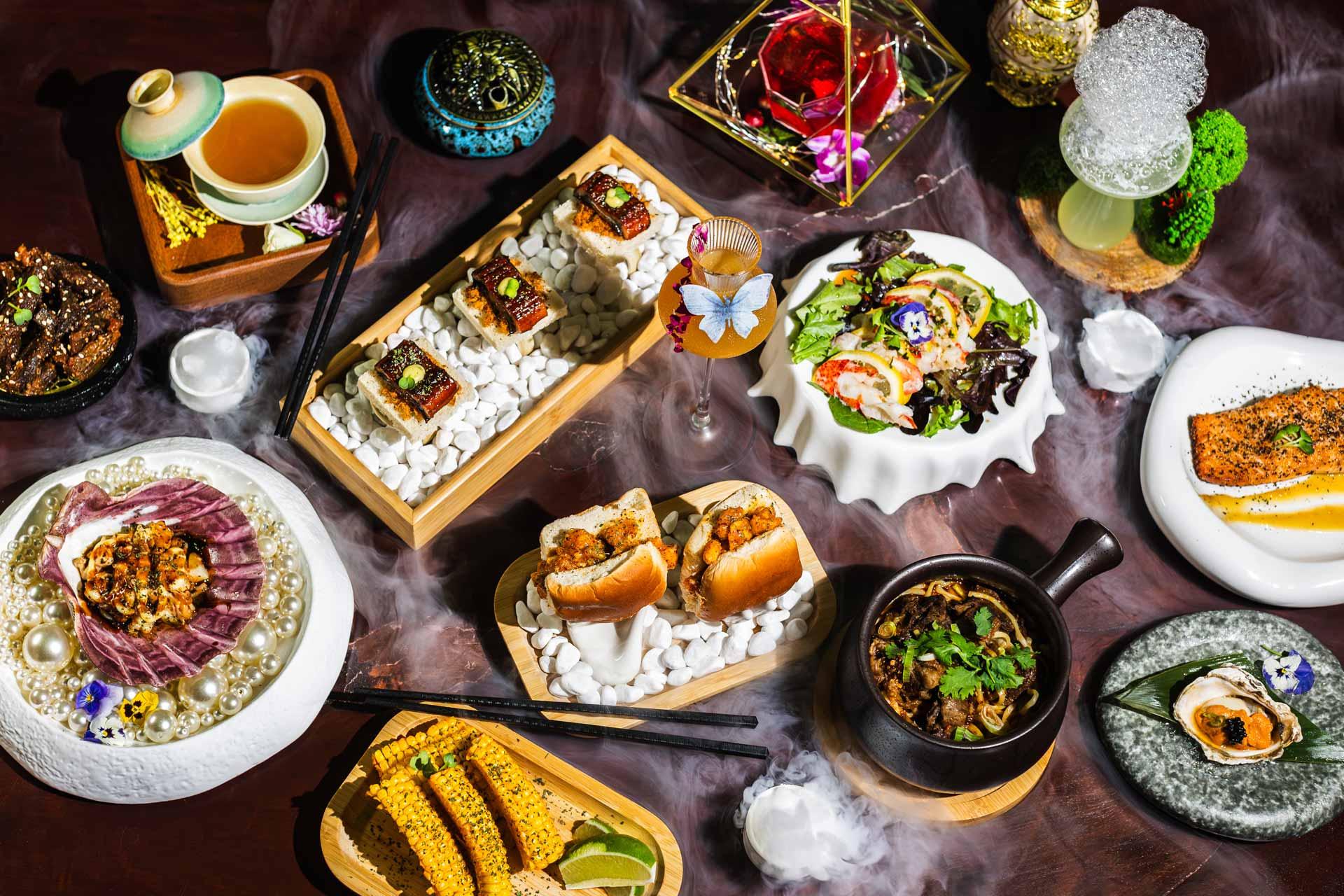 An assortment of gourmet dishes on a table includes sushi, a salad, a steak, corn on the cob, sliders, and various small side dishes. The presentation features artistic plating with decorative elements and dry ice for a smoky effect.