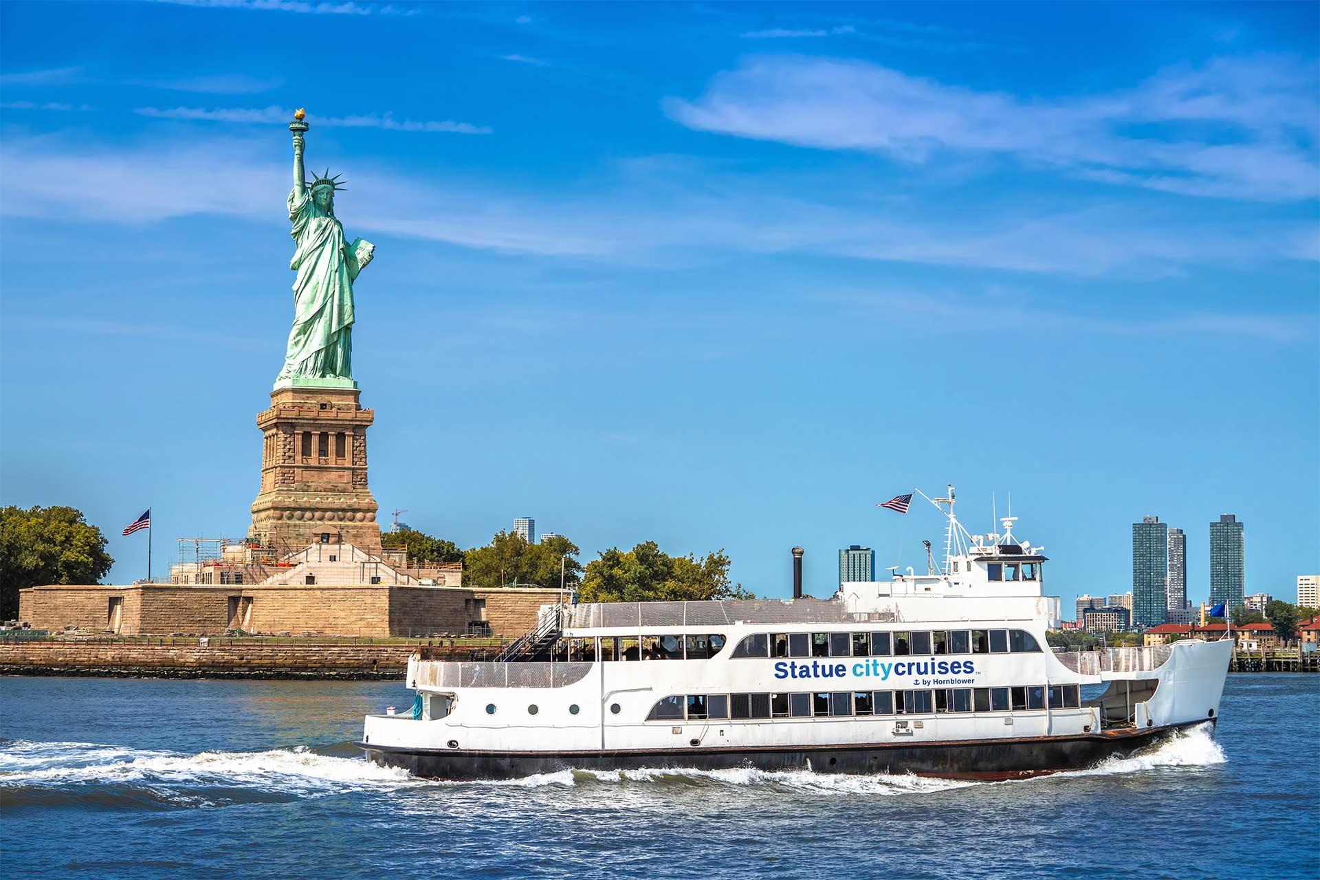 A white ferry labeled "Statue City Cruises" sails on the water near the Statue of Liberty, with a clear blue sky and city skyline in the background.