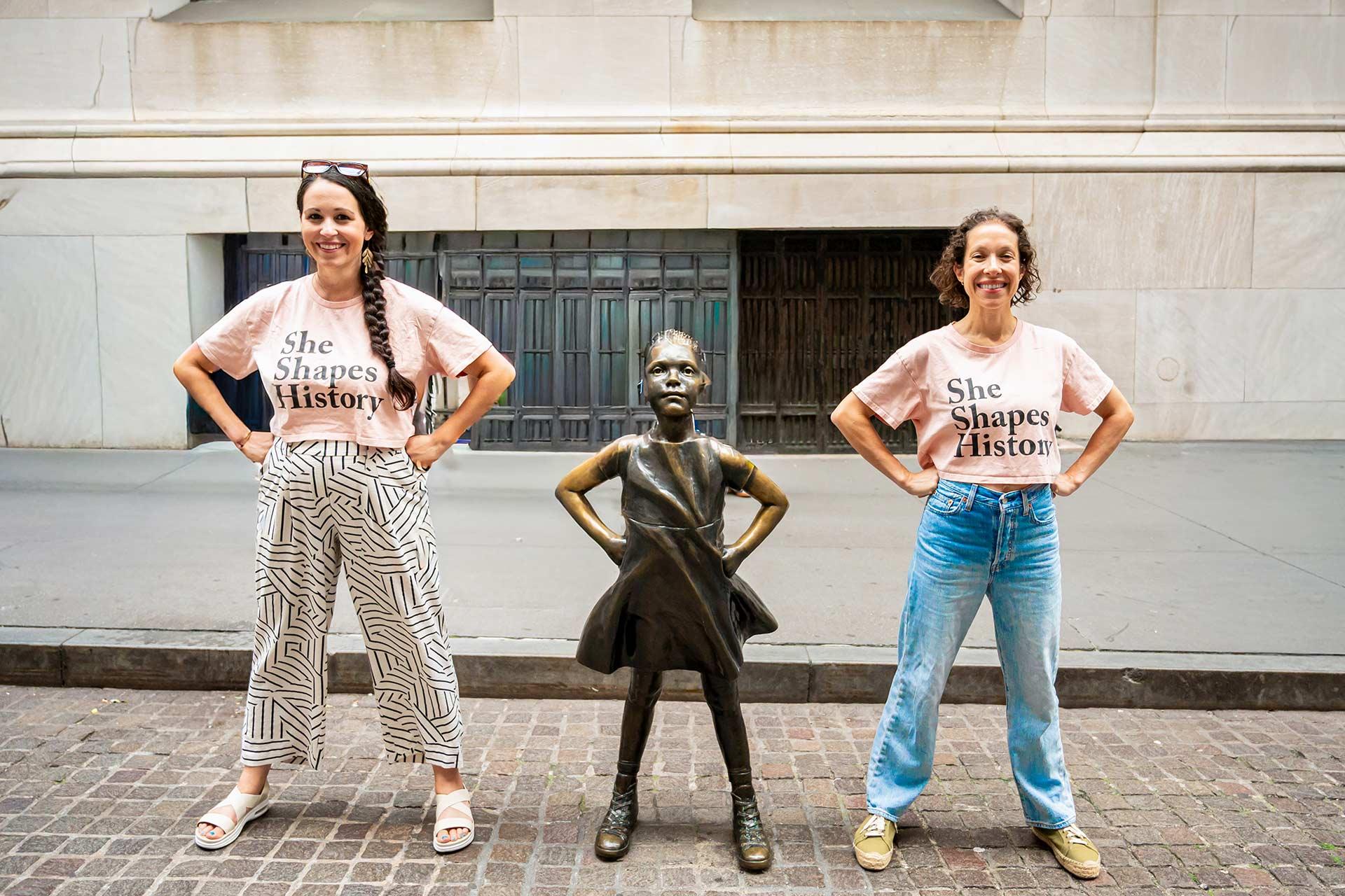 Two women stand on either side of the "Fearless Girl" statue, mimicking its pose. They wear matching "She Shapes History" shirts and smile, standing on a city sidewalk in front of a large building.